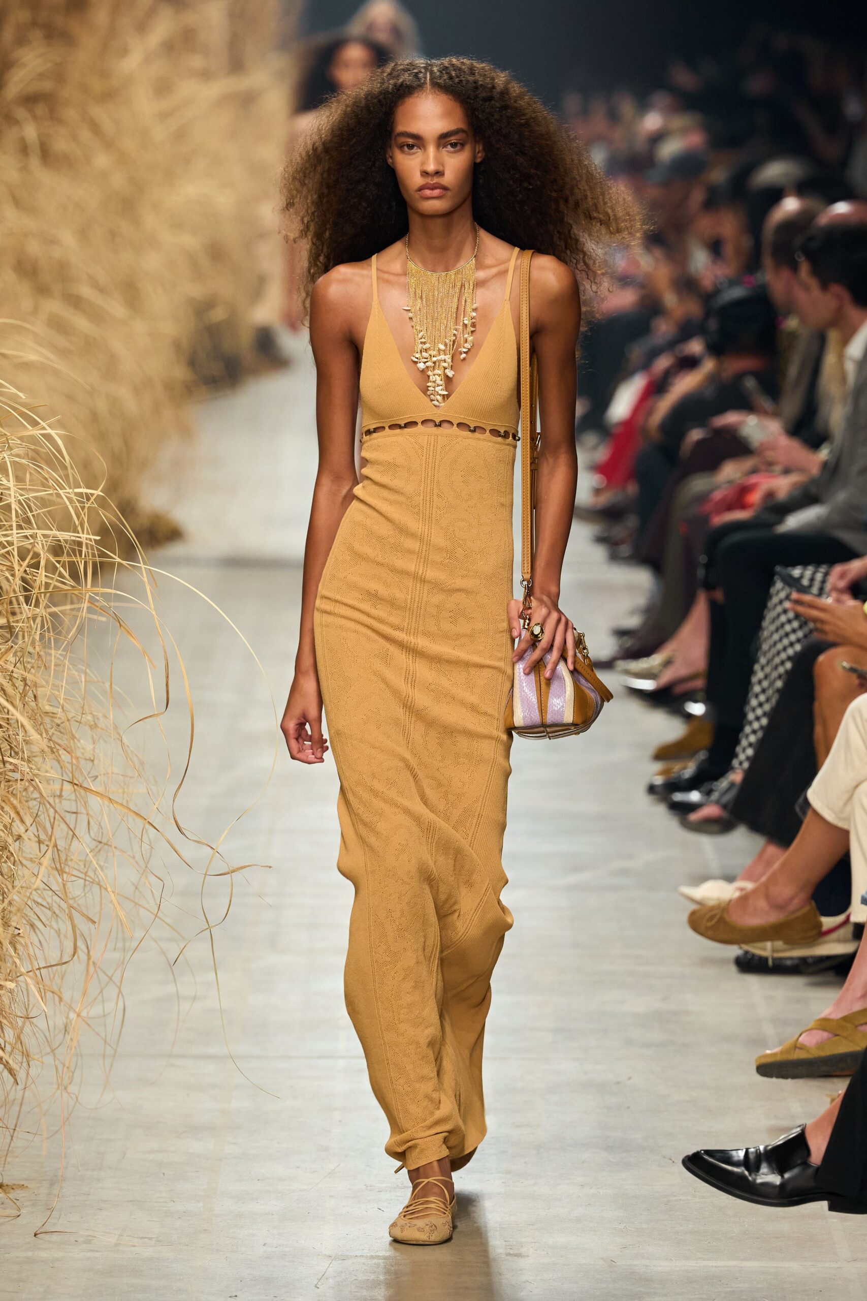 Gold fringe bib threaded with shell drops over the sternum. Ochre knit column dress, plunge bodice cut by a ringed underbust seam. Striped mini shoulder bag with tan leather piping. Lace-up almond-toe flats in matching pointelle knit. Walking back from the beach bar with salt drying on your collarbones.