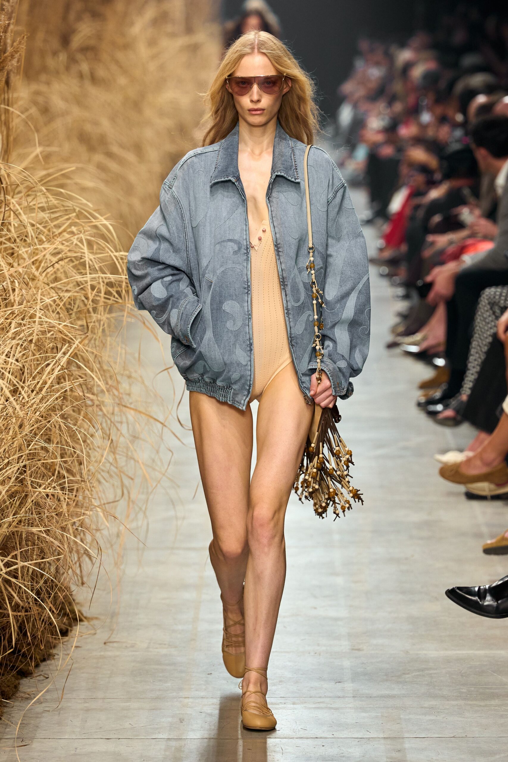 Amber shield sunglasses with a rimless brow. Oversized faded-denim bomber with tonal scroll embroidery and gathered hem. Nude pointelle bodysuit, a few bead drops at the deep V. Brown tassel bag strung with bone and amber beads. Tan leather wrap flats. Walking back from the dunes with sand still inside your shoes.