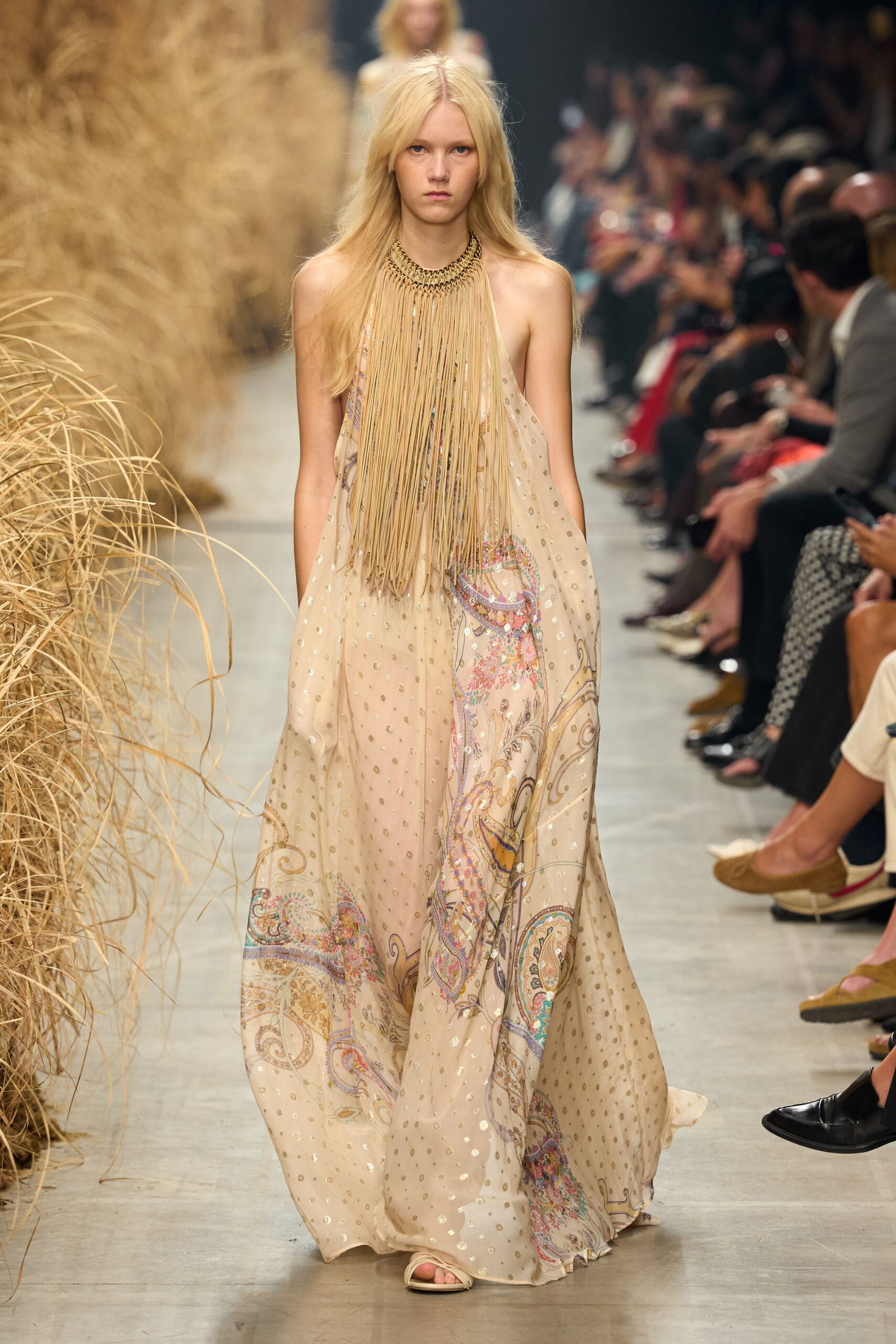 Bib necklace of long sand suede fringe from a braided collar. Blush chiffon halter gown, scattered gold foil dots and paisley border print. Flat ivory sandals, barely-there straps. Walking through dry grass after a wedding that ran into sunset.