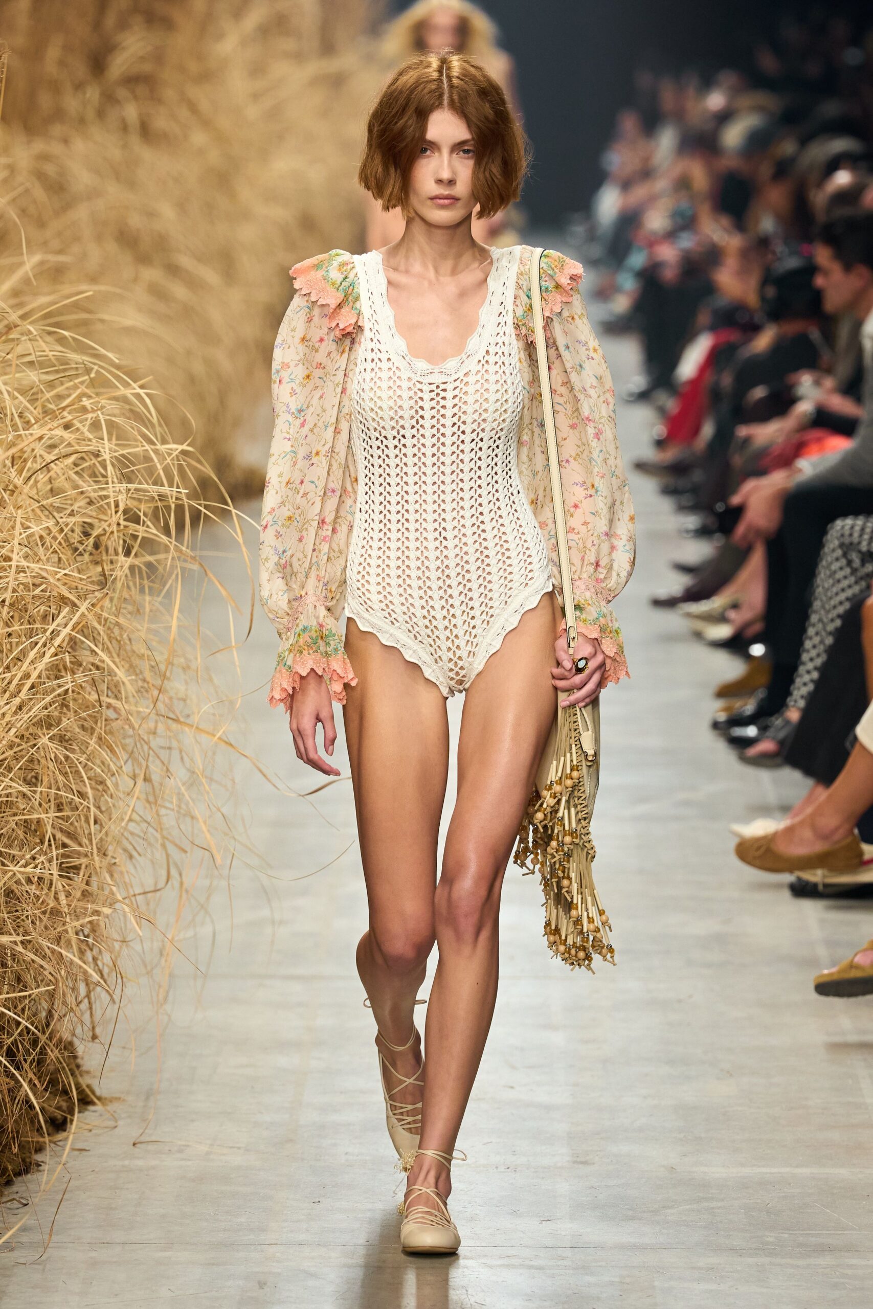Openwork cream crochet bodysuit with a scalloped leg line. Sheer floral chiffon blouse, peach crochet ruffles at shoulder and cuff. Ivory shoulder bag with wicker panel and swinging bead fringe. Nude lace-up flats, ties winding up the ankle. Walking back from a dune cottage with sand still stuck to your calves.