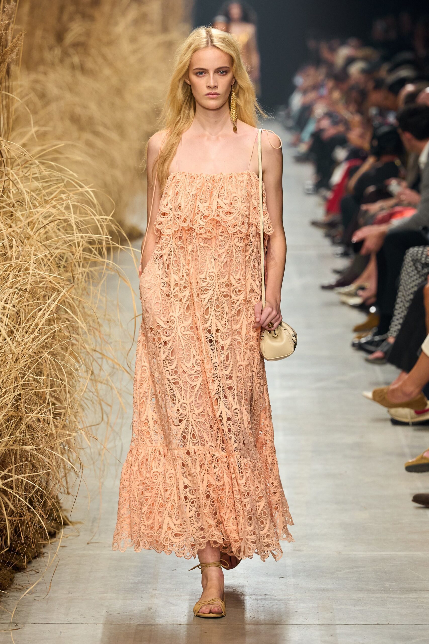 Blush guipure lace slip dress with a loose flounce across the bust. Single chain tassel earring brushing the shoulder. Cream drawstring pouch on a narrow strap. Tan wraparound flat sandals, ties winding the ankle. Walking back from a beach wedding after the light has turned honey.