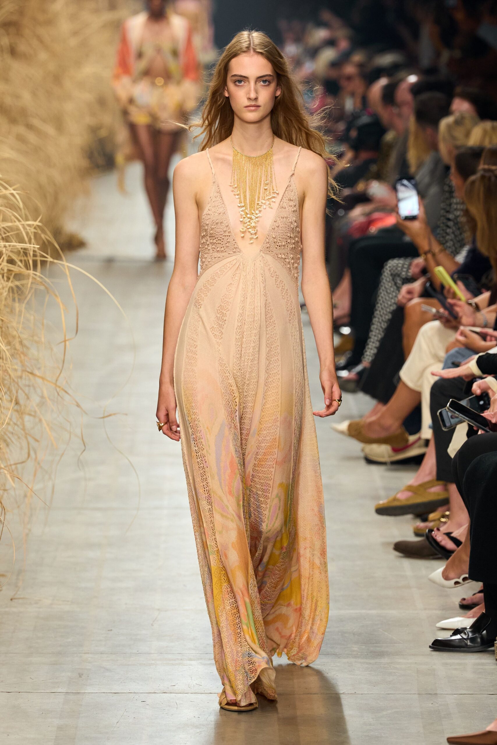 Long gold fringe necklace tipped with clustered shell-like beads. Blush plunge dress in openwork knit, watercolor panels pooling at the hem. Flat nude sandals barely visible beneath the dragging skirt. Slim gold rings on two fingers. Walking back from the beach bar with sand still stuck to your ankles.
