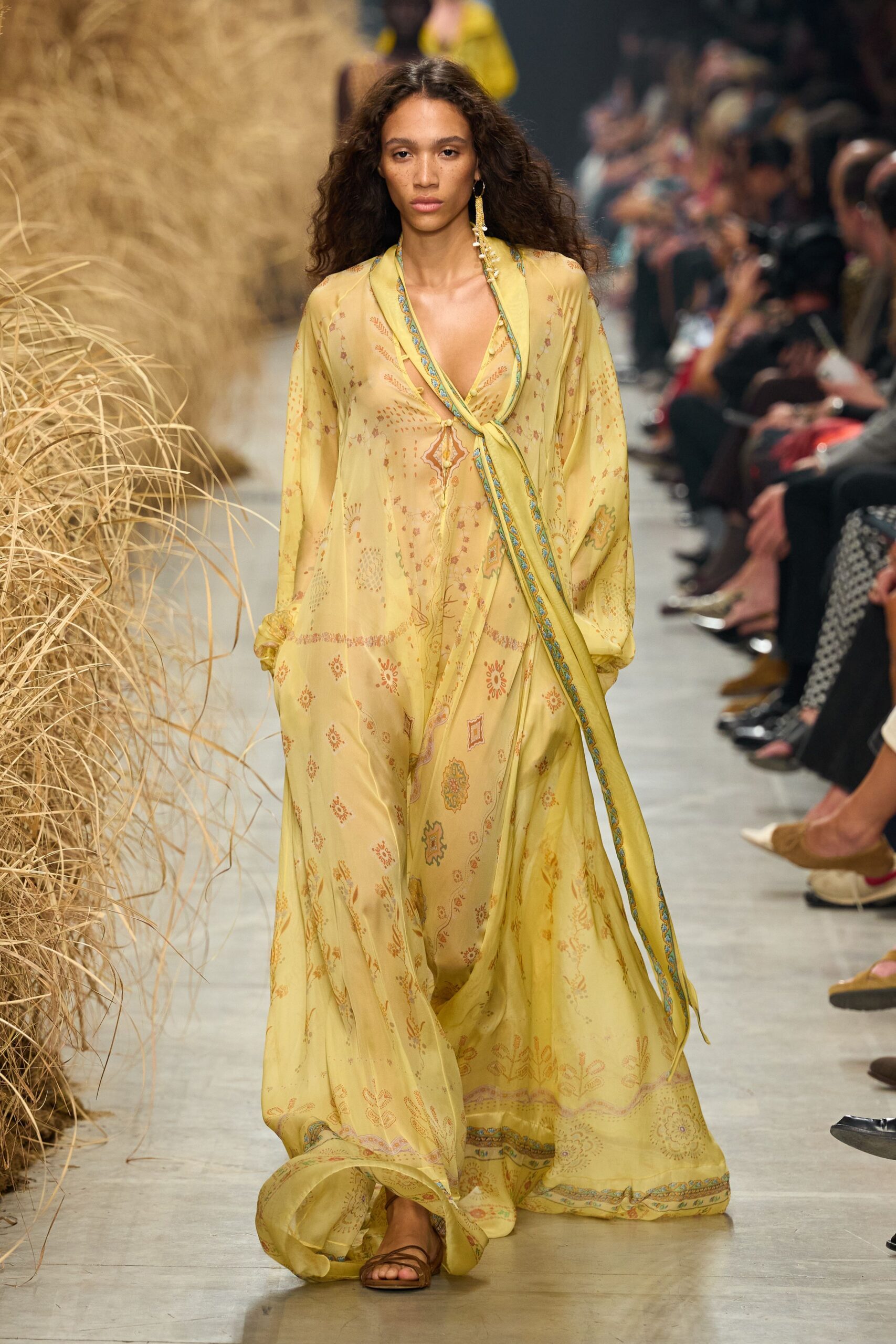 Butter-yellow silk caftan, sheer and scattered with faded tile-print motifs. Turquoise-bordered ties loop loosely across the plunging front. Single tassel earring with dangling beads. Flat brown leather thong sandals. Walking out to breakfast at a desert lodge before the heat starts.