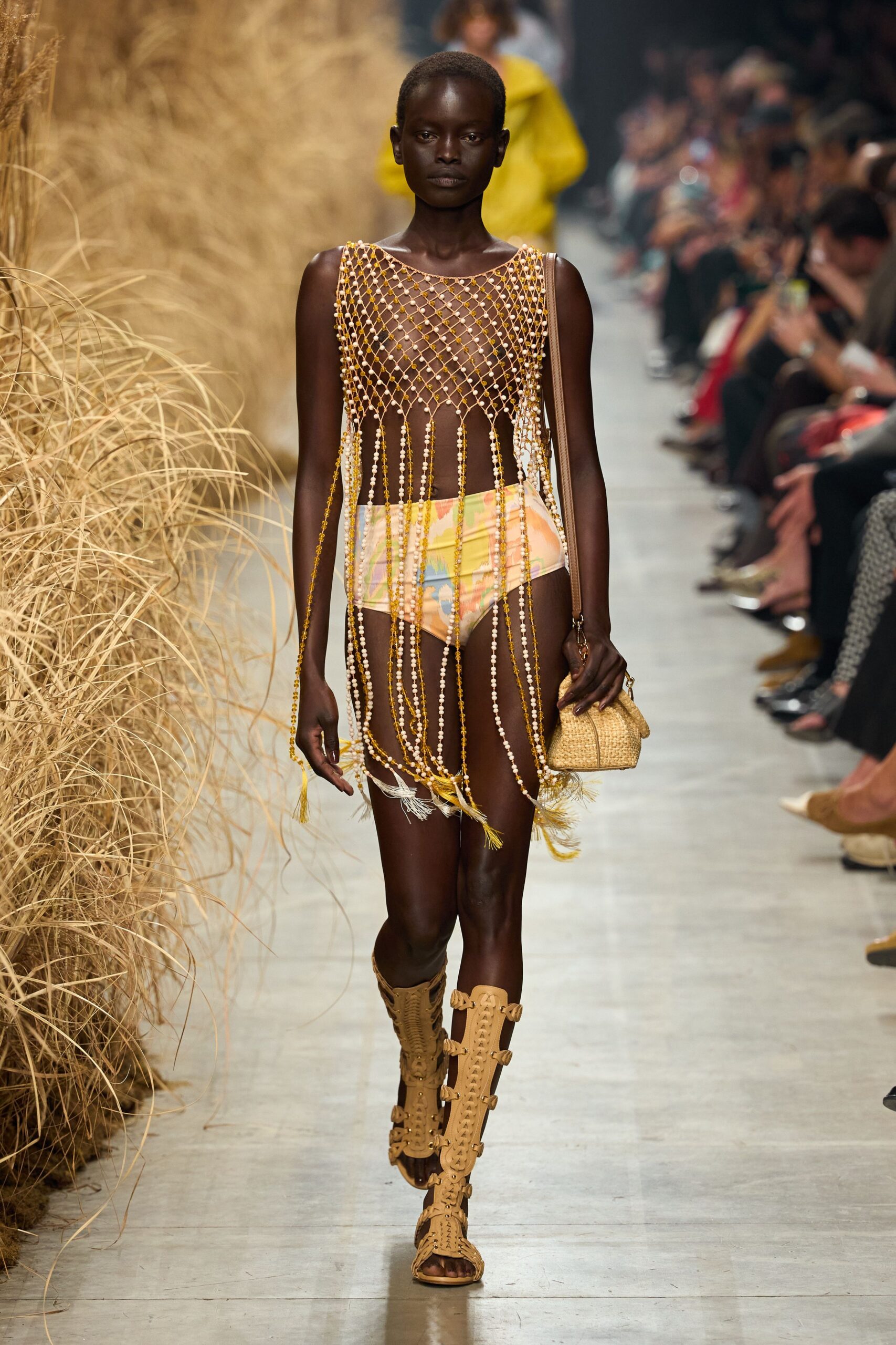 Amber and ivory beaded mesh, tasselled strands falling past the hips. Pastel printed briefs cut high on the leg. Mini raffia bag on a narrow tan leather strap. Knee-high tan leather sandals, laddered with cutout loops. Walking back from the beach bar with salt still drying on your skin.