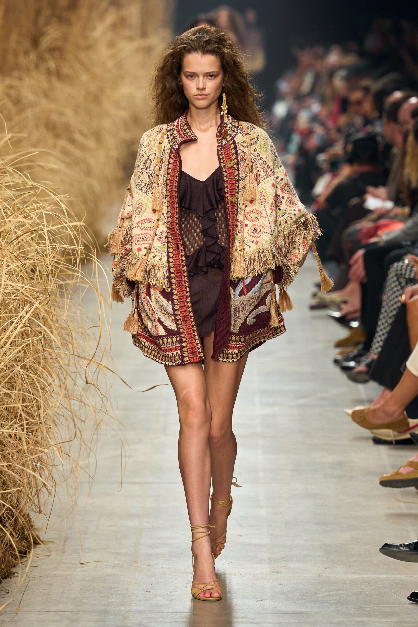 Cream jacquard cardigan with burgundy borders and thick swingy fringe. Deep brown mini dress, ruffled plunge over an open ladder-knit panel. Single tasseled drop earring brushing the collar. Tan wrap sandals laced high at the ankle. Walking back from a dune picnic with burrs caught in the hem.