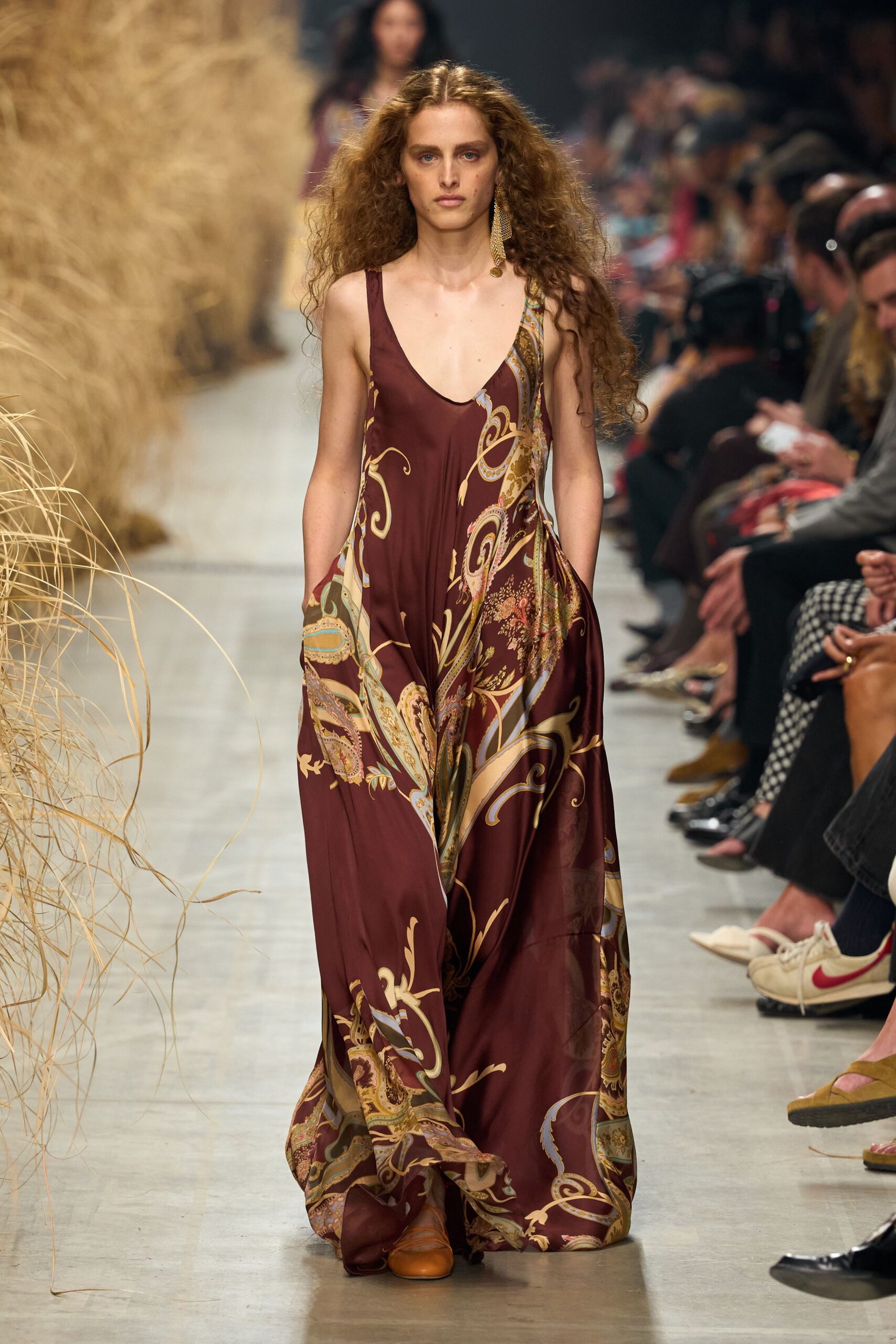 Wine satin maxi dress with scarf-print panels and deep scooped neck. Single gold tassel earring brushing the shoulder. Tan leather flats peeking from the pooled hem. Walking through a dry conservatory with dust on your ankles.