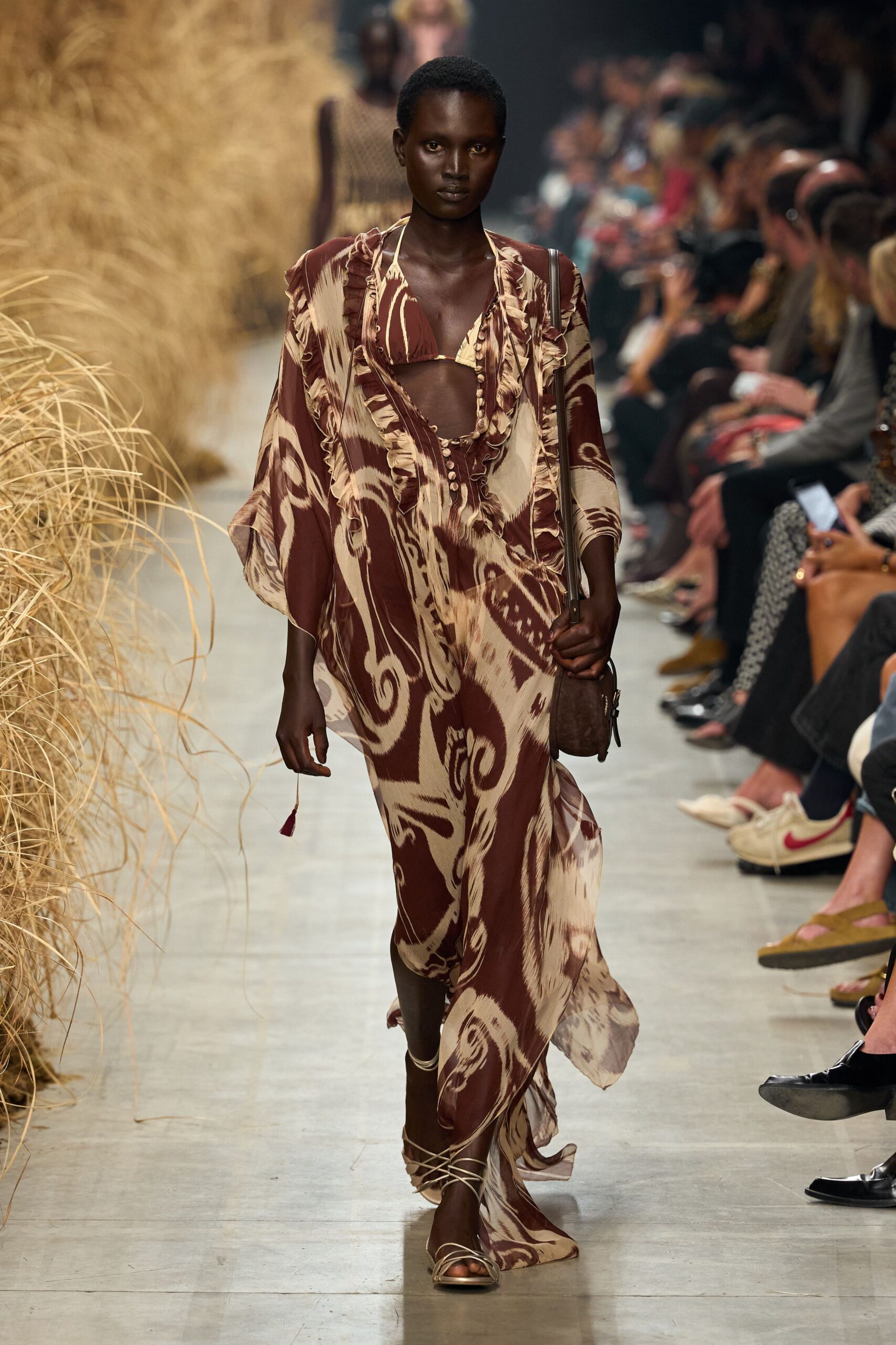 Chestnut-and-cream swirled chiffon caftan, ruffled edges framing a plunging opening. Matching triangle bikini top tied on with thin cream cords. Narrow brown suede pouch hanging low on a long strap. Cream wraparound laces spiraling over flat leather soles. Walking back from the beach bar with salt drying on your shoulders.