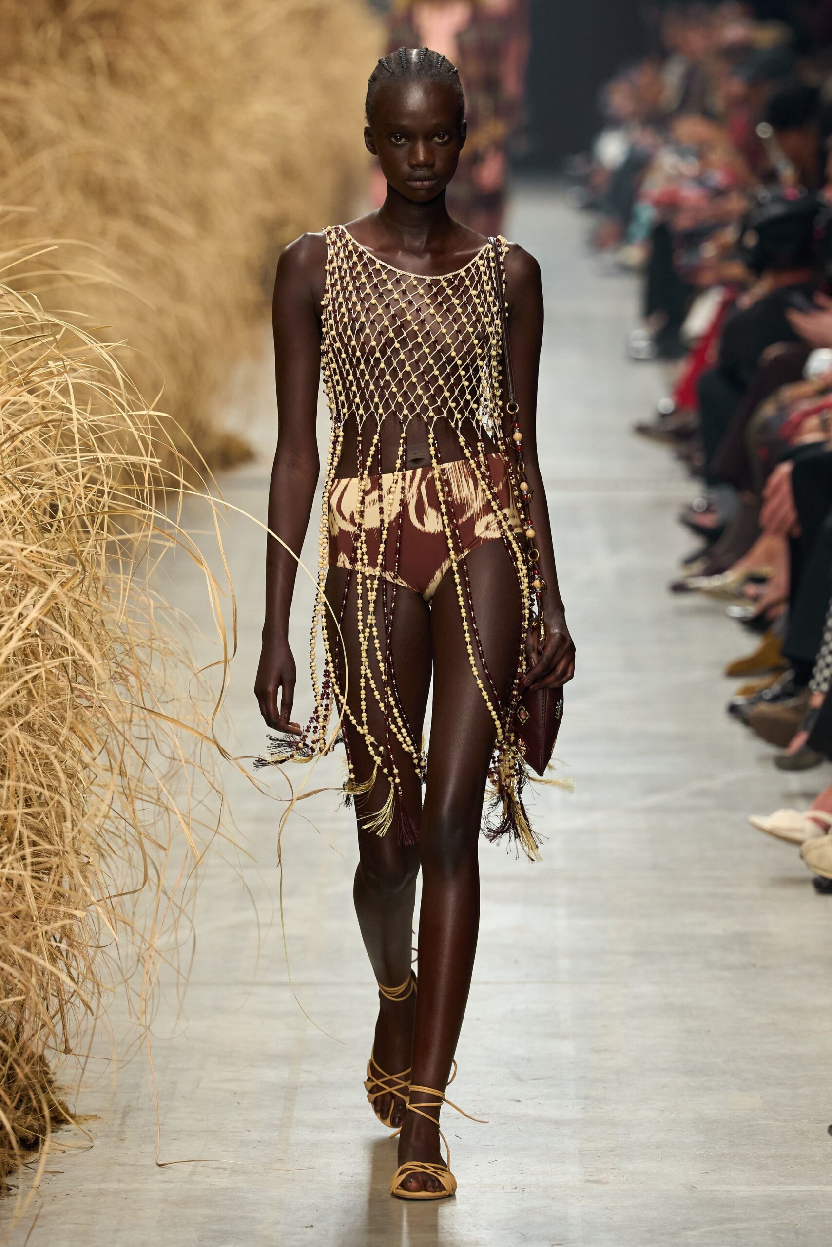 Cream and oxblood bead lattice, long tassels swinging below the hip. Brown-and-cream ikat briefs cut high on the thigh. Burgundy leather pouch with beads threaded down the strap. Tan wrap sandals laced thin around the ankle. Walking back from a beach bonfire with ash still in the air.