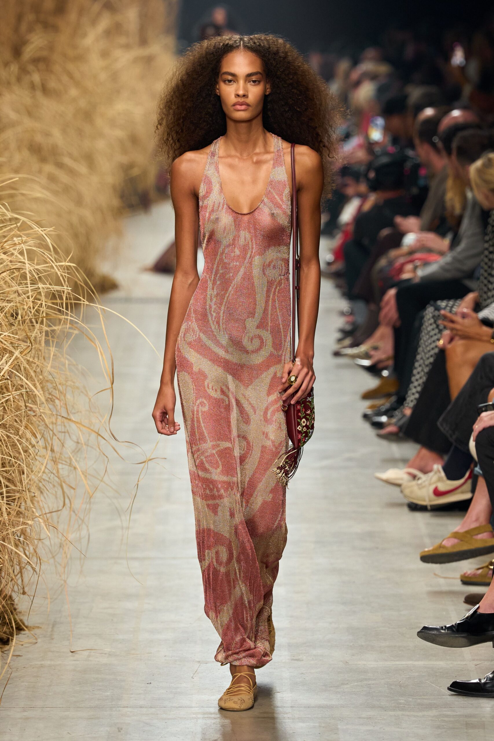 Rose and sand metallic knit column dress with a low scooped racer front. Burgundy tapestry pouch with beaded fringe brushing the hip. Tan wrap-tie flats, toes stitched with a tiny floral motif. Large oval ring on the right hand. Walking back from a seaside wedding after the band has packed up.