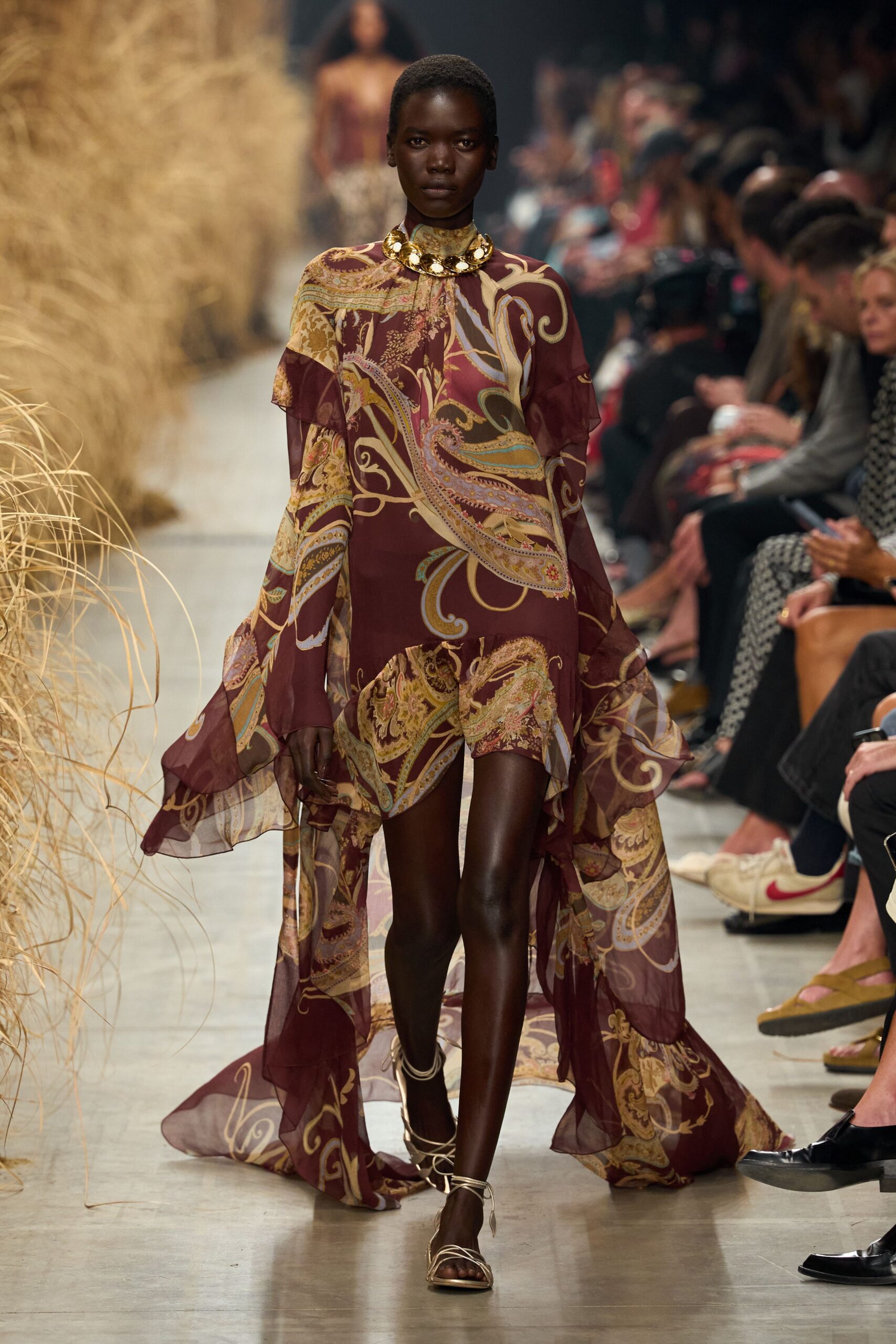 Hammered gold disc collar sitting tight above the clavicle. Burgundy sheer chiffon dress in sprawling paisley, with fluted sleeves and a trailing waterfall hem. Ivory lace-up sandals, cords winding twice around the ankle. Walking past dry grass at a vineyard wedding after the sun drops.