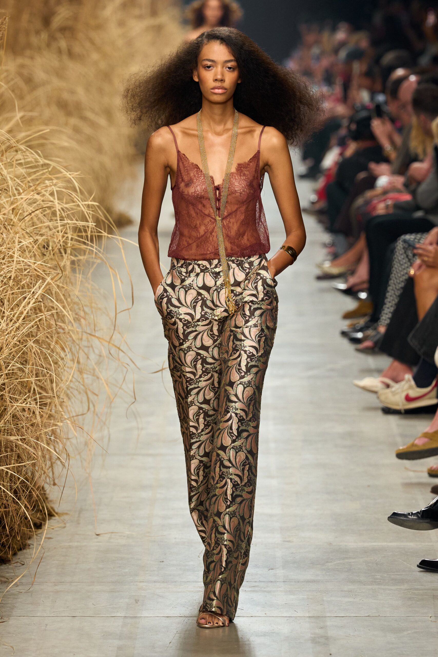 Sheer claret lace cami with a scalloped V and tiny center lacing. Long gold mesh necklace ending in a tassel past the waist. Wide gold cuff. High-waisted brocade trousers in blush and olive arabesques. Barely there metallic sandals. Walking through a botanic glasshouse after the last wedding guest leaves.
