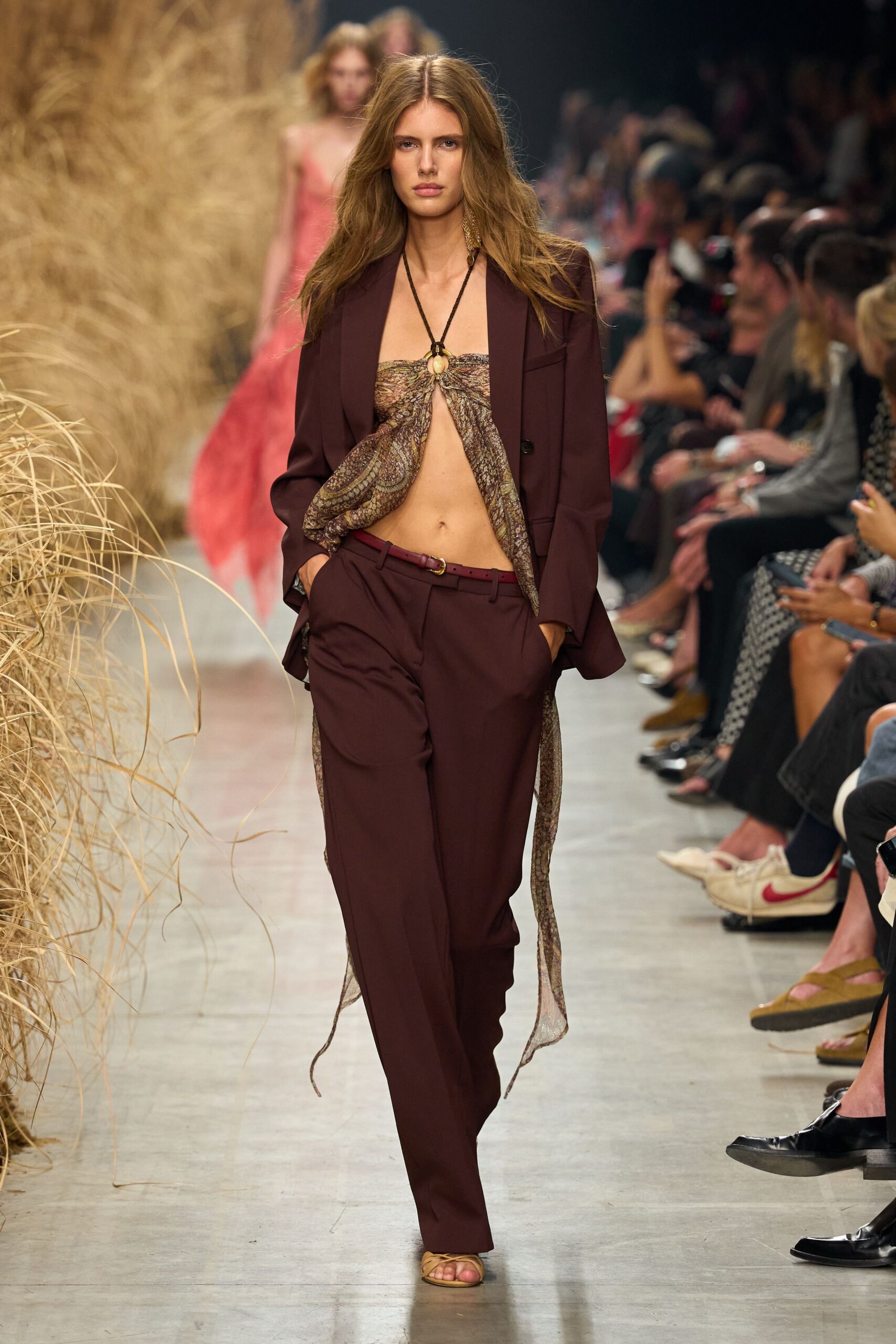 Deep plum blazer in dry suiting, cut loose through the body. Paisley chiffon halter knotted through a horn ring, hem streaming long. Matching straight trousers with a low rise and soft break. Narrow oxblood belt with a small brass buckle. Tan leather thong sandals. Walking back from a vineyard lunch with dust on your ankles.