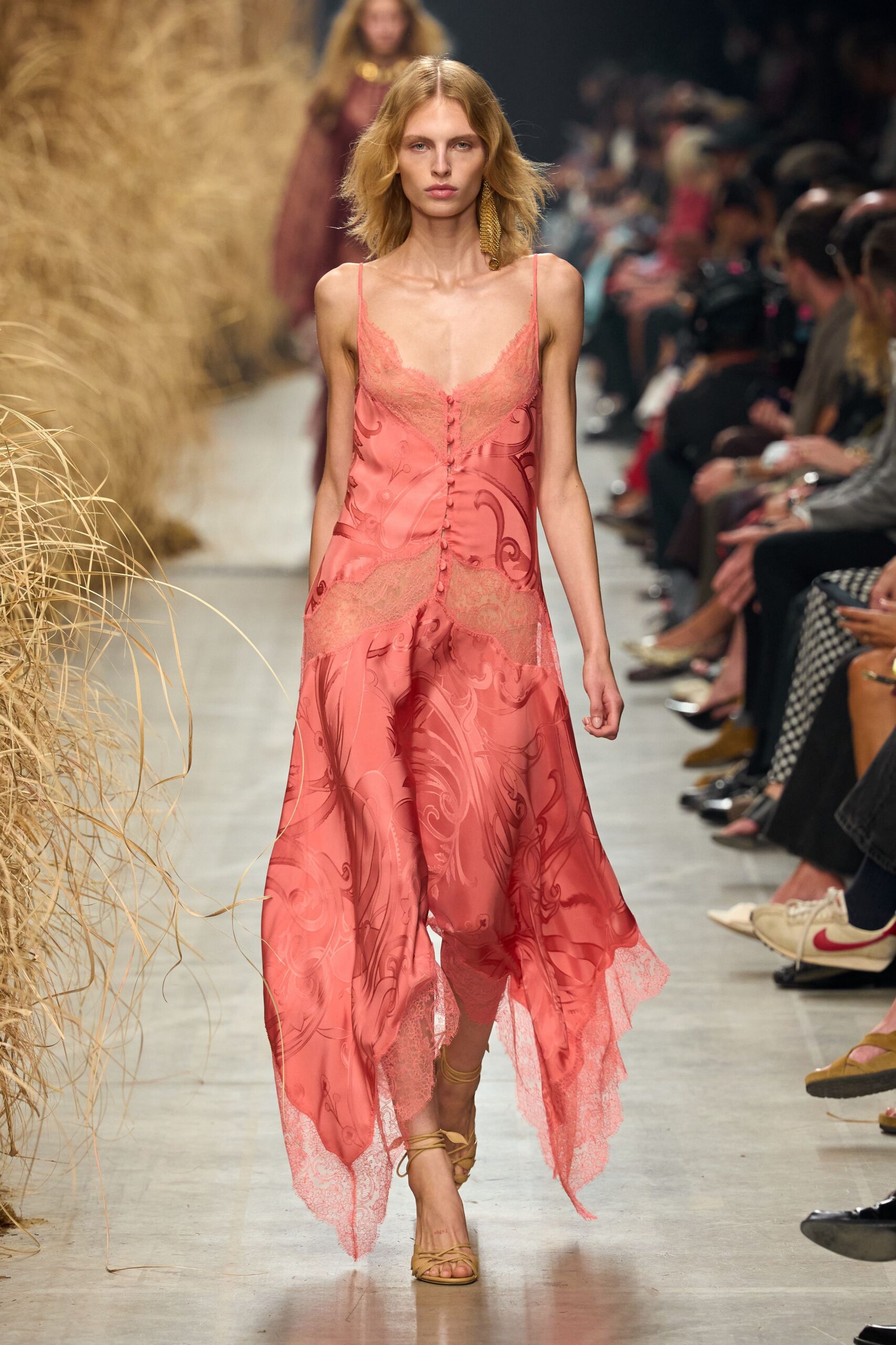 Coral satin slip dress with lace insets, rouleau buttons, and a handkerchief hem. Single gold tassel earring brushing the shoulder. Tan wrap sandals laced slim around the ankle. Walking back from a beach wedding with dune grass still catching the light.