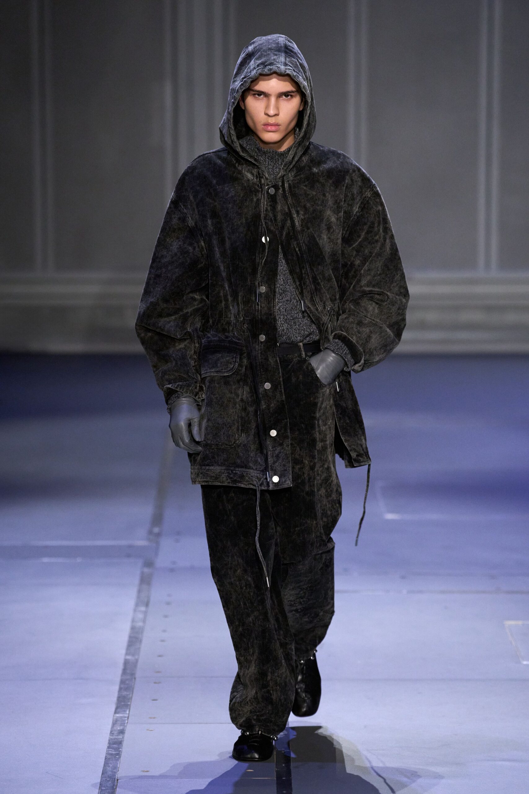 Washed black corduroy hooded jacket with silver snaps and dangling drawcord ends. Charcoal marled knit sweater, collar hugging high under the hood. Matching corduroy trousers falling loose over the shoes. Smooth grey leather gloves. Black leather shoes with rounded toes. For crossing a ferry deck in weather that keeps changing.