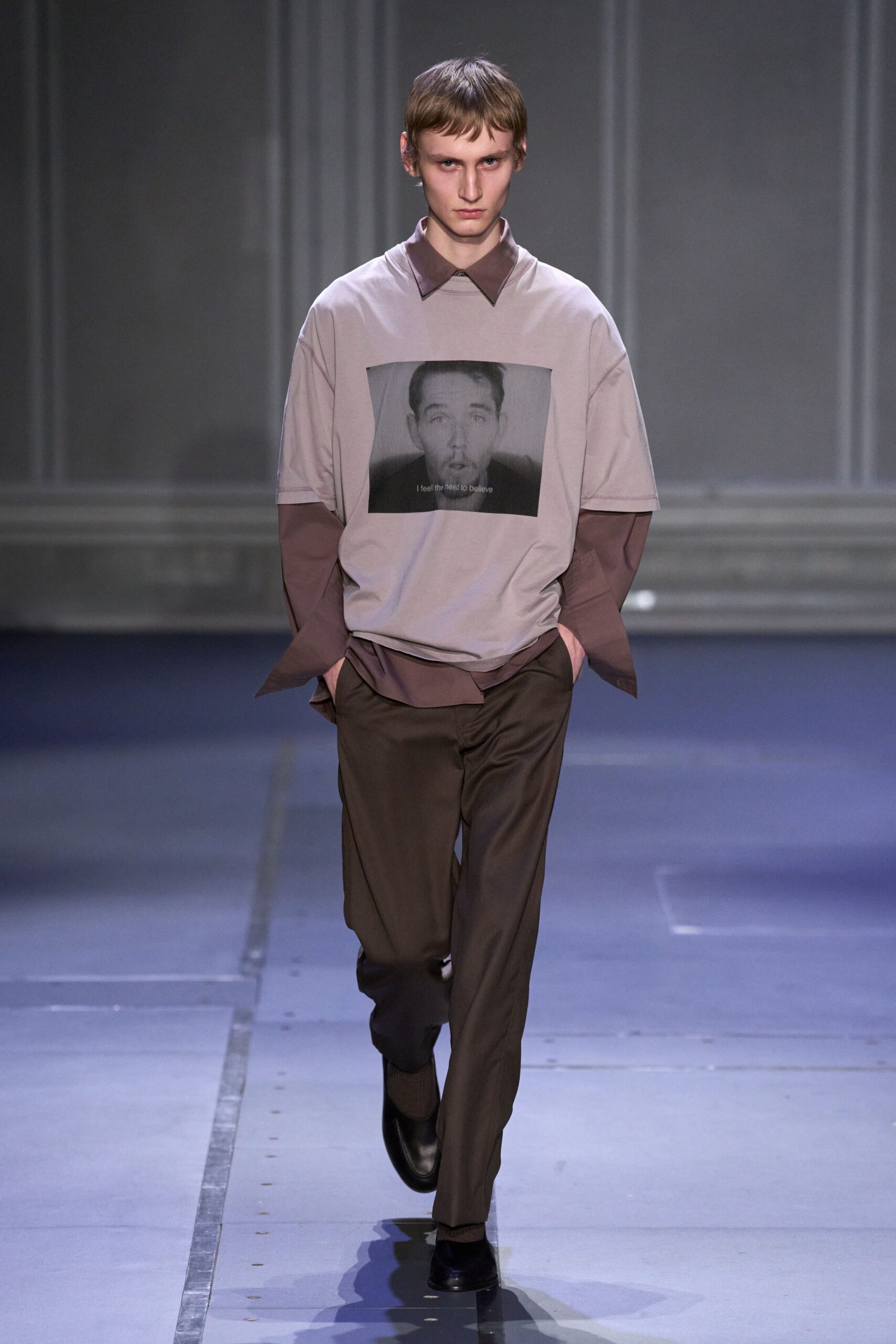 Pale greige jersey tee with a grayscale face print centered wide. Mauve button-up layered underneath, long cuffs hanging past the hands. Dark brown tailored trousers with a clean flat front. Black leather slip-ons with a blunt square toe. Walking empty museum corridors after the audio guide has cut out.