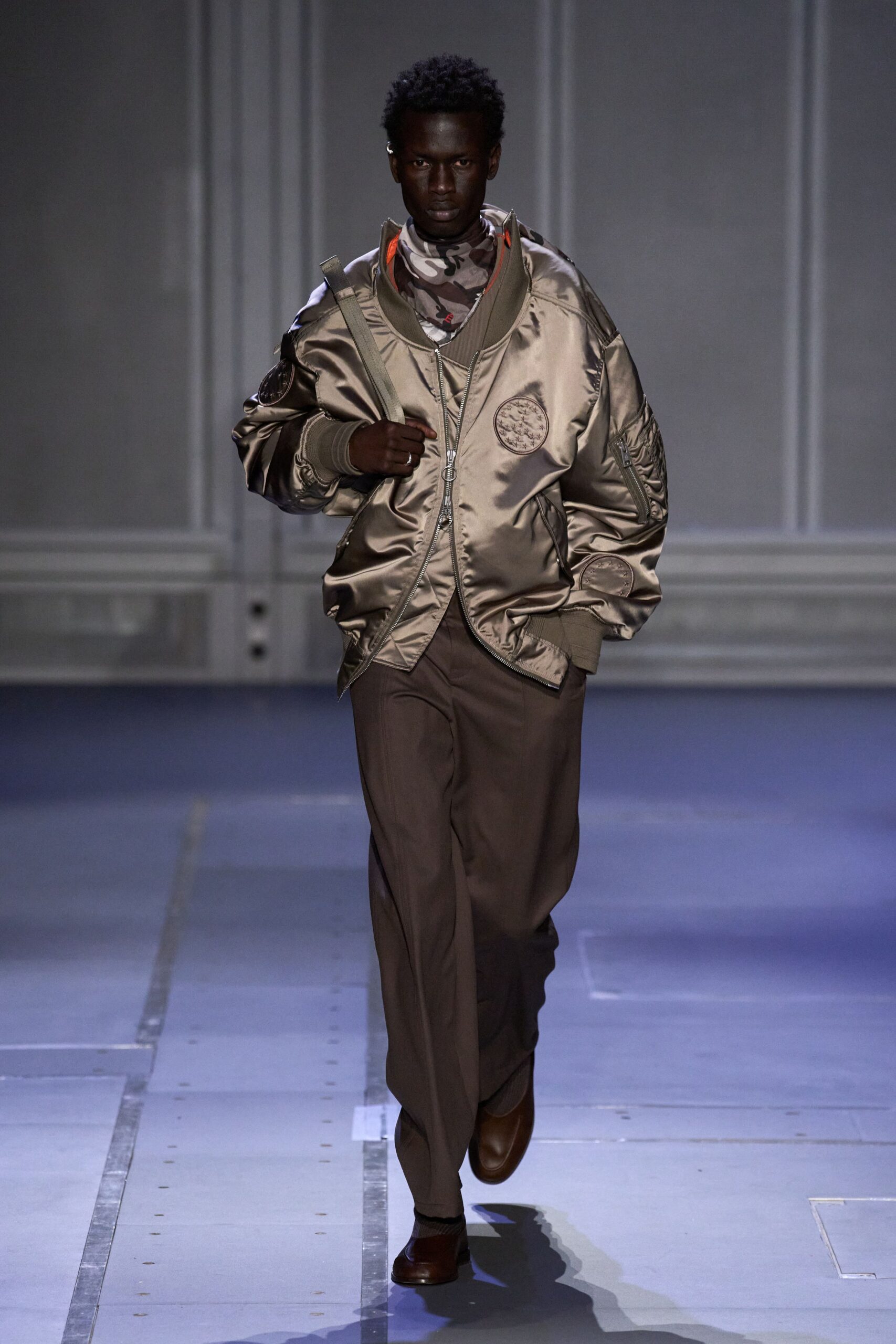 Champagne satin bomber with orange lining flashing inside the dropped collar. Brown camouflage mesh turtleneck, close at the throat. Dark cocoa trousers with a soft break over the shoe. Chestnut leather square toes. Webbing strap slung over one shoulder. Whatever you wear home after missing the last military shuttle.