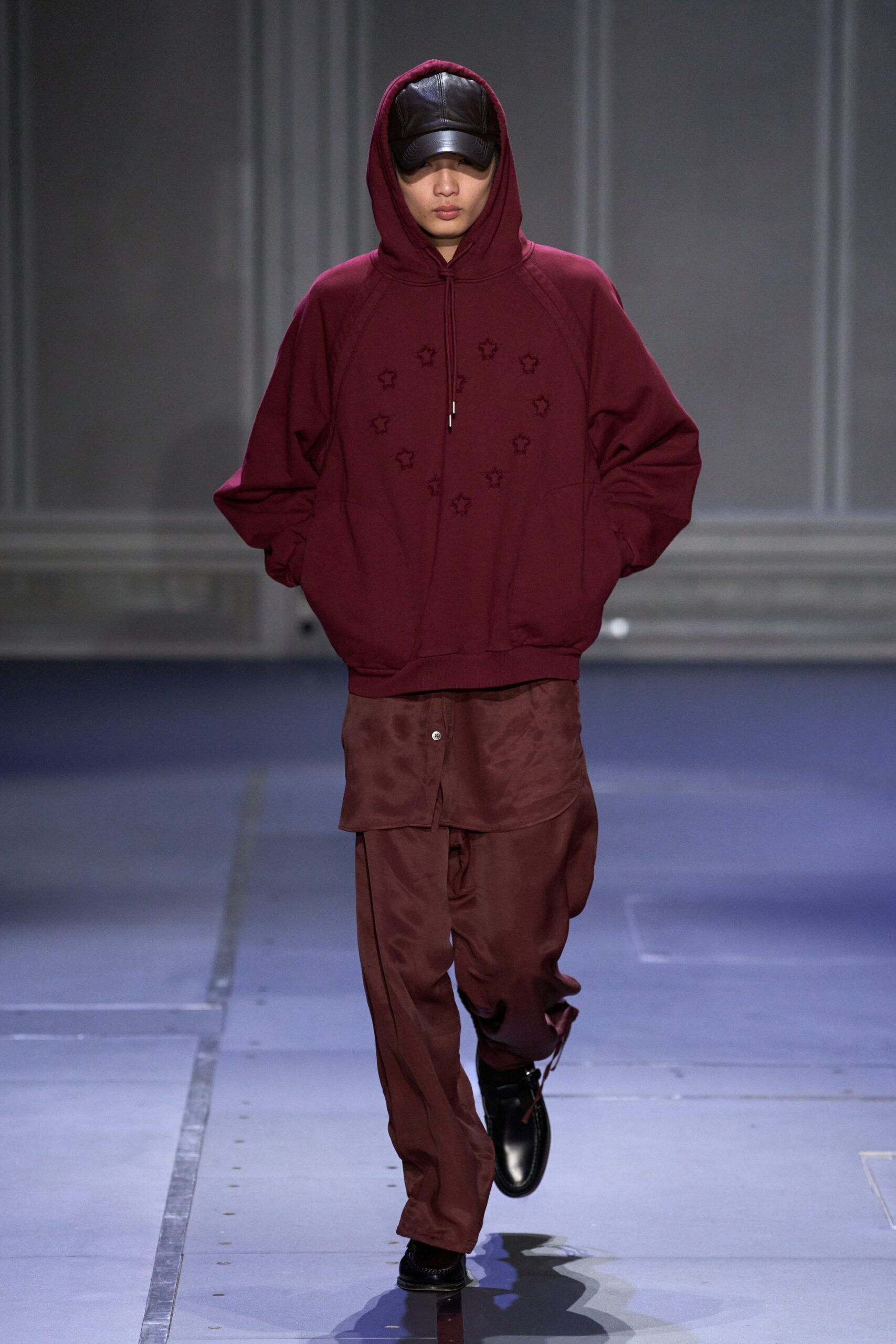 Black leather cap with a deeply curved brim. Burgundy fleece hoodie, star cutouts ringed across the chest. Mahogany satin shirt hanging below the sweatshirt hem. Loose brown trousers with a drawcord flicking at the ankle. Black leather slip-ons, blunt and glossy. Walking to the corner store just after rain, hood still up.