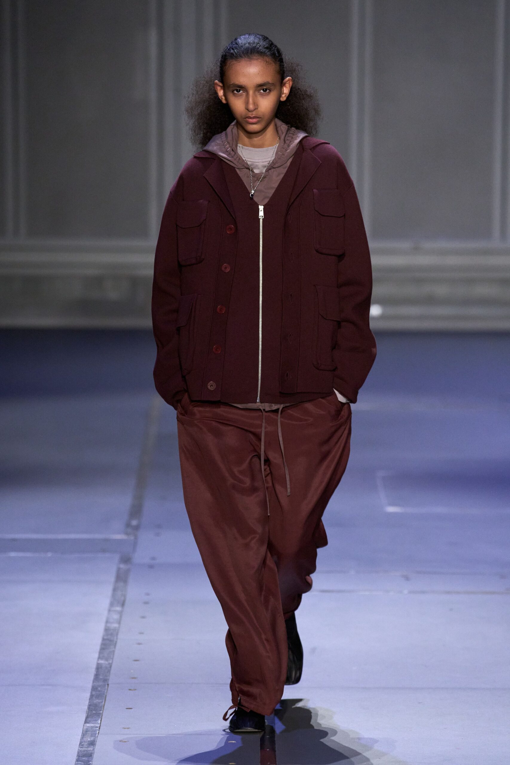 Oxblood wool jacket with four flap pockets and matte horn buttons. Mauve nylon hoodie peeking out, cords hanging past the fly. Pale taupe crewneck tee at the collar. Cinnamon satin trousers, loose through the leg. Black square-toe boots. For crossing the station concourse before dawn with coffee going cold.
