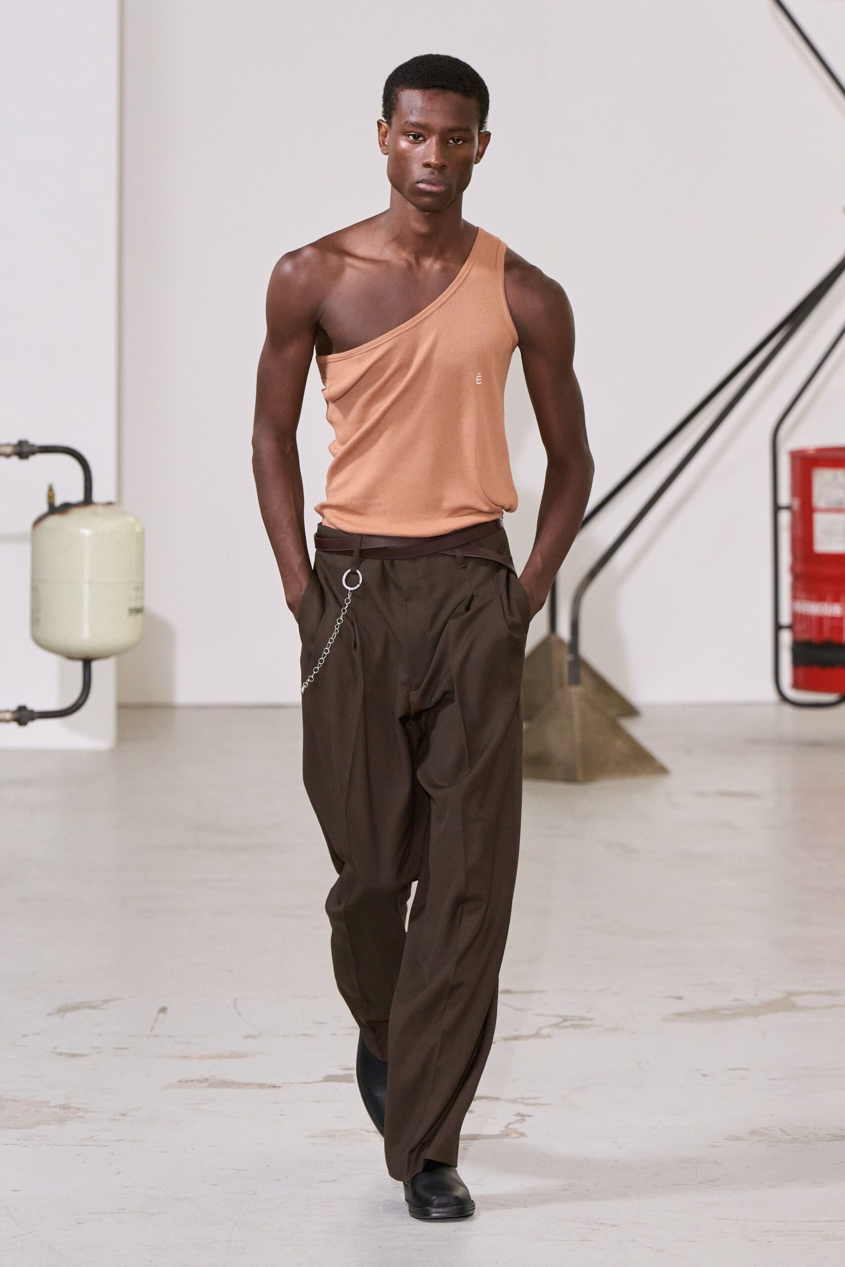 Peach ribbed tank cut on a steep one-shoulder slant. Dark chocolate trousers with a dropped wrap waistband and deep pleats. Silver curb chain hooked to a single oversized ring. Square-toe black leather boots. Crossing an empty design studio after someone left the fire door open.