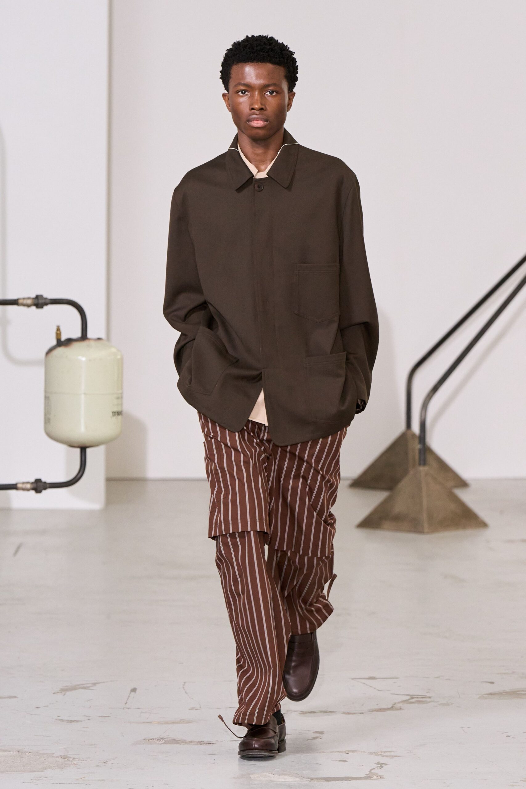 Dark brown work jacket with oversized patch pockets and one visible top button. Cream crewneck peeking out in a narrow uneven strip. Chestnut trousers in white banker stripes, extra panel hanging over one thigh. Square-toe brown leather slip-ons. Crossing a school gym converted into a craft fair before the doors open.