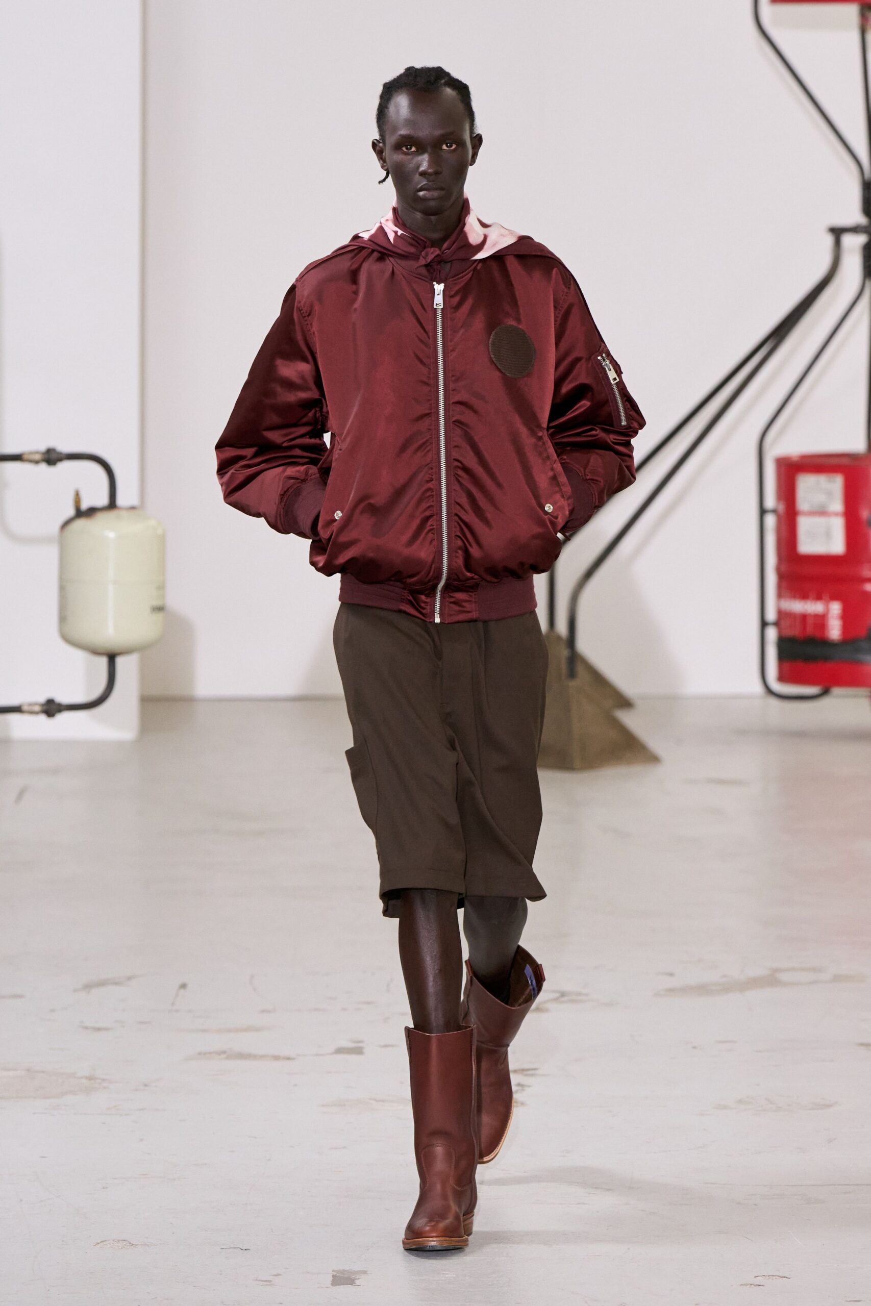 Burgundy satin bomber, ruched at the zip with a sleeve utility pocket. Pale pink lining peeking from the hood knotted at the throat. Dark brown tailored shorts, pressed front falling just below the knee. Chestnut leather mid-calf boots with a squared apron toe. Crossing an empty ferry terminal before the ticket windows open.