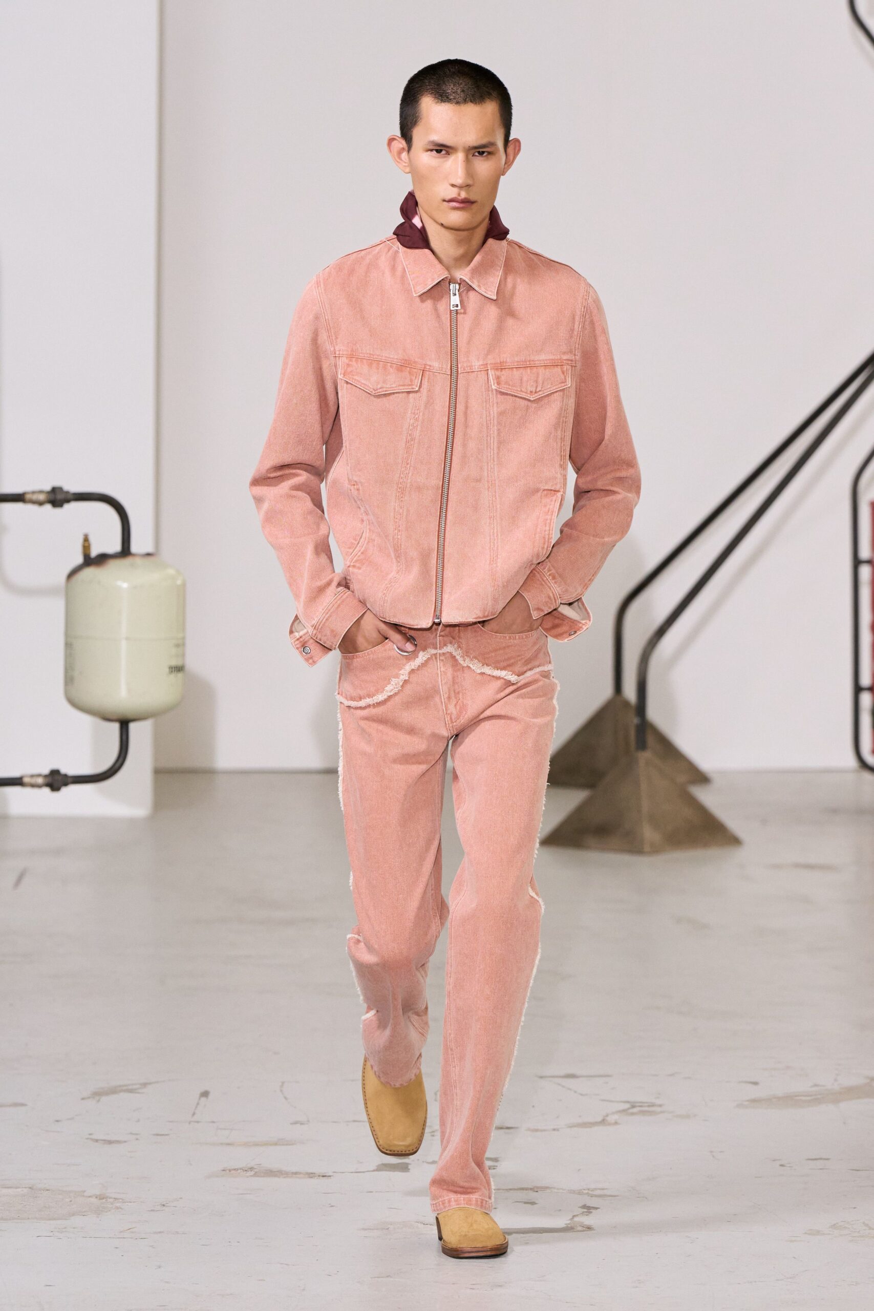 Burgundy shirt collar popped high behind the jacket neck. Dusty rose zip jacket in washed denim, flap pockets sharp at the chest. Matching pink jeans with frayed cream seam strips snaking over the hips. Square-toe sand suede boots. Walking through a converted warehouse before the paint smell has left.