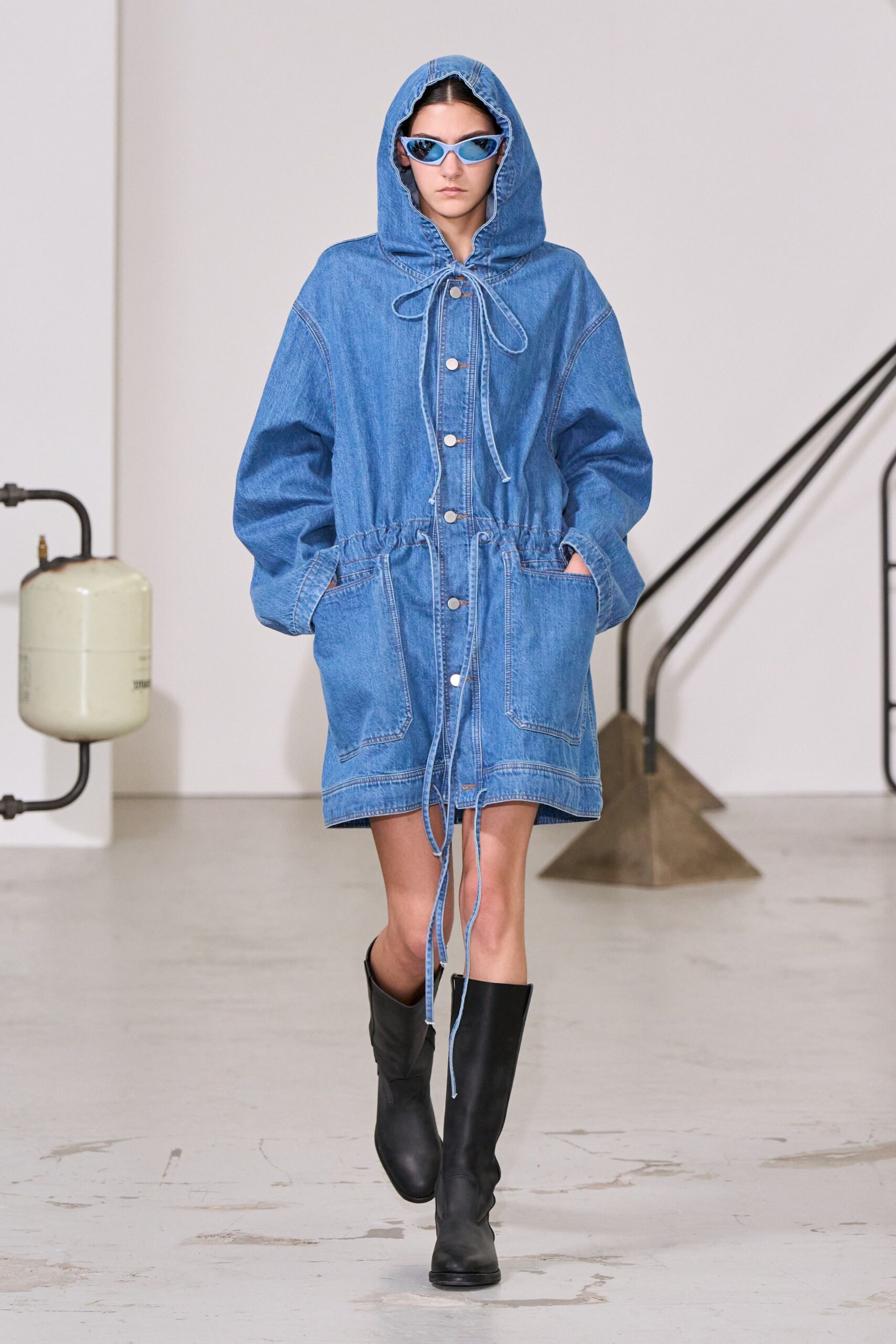 Powder-blue cat-eye frames with mirrored lenses. Washed indigo denim parka, hood cinched with long fraying cords and white snaps. Black knee-high boots with blunt round toes. Walking across a concrete courtyard before the rain starts.
