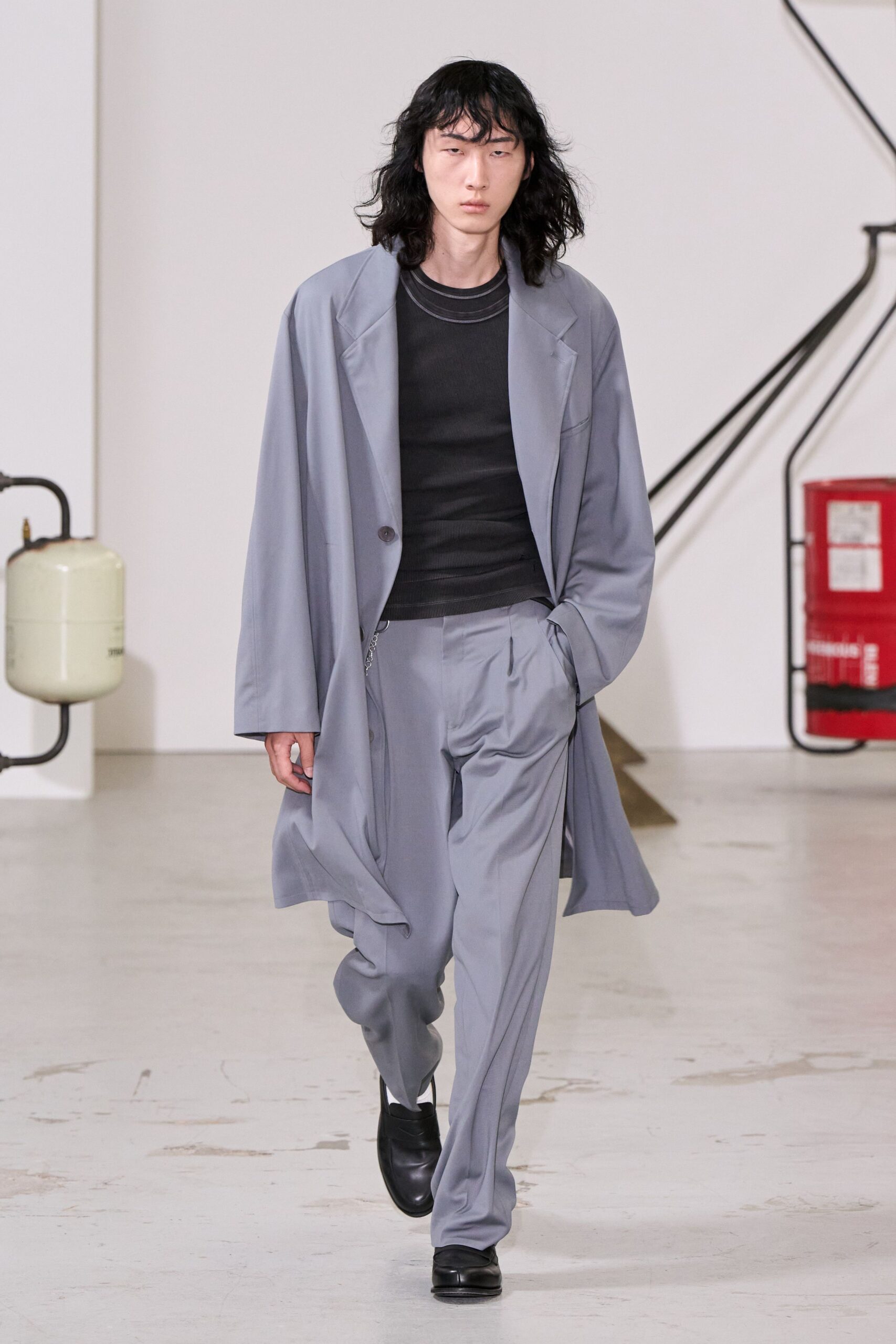Slate draped overcoat in fluid suiting, one dark button set low. Black ribbed tank with a double-bound crewneck tracing the collarbone. Matching slate trousers, softly pleated and breaking over the shoe. Square-toe black leather slip-ons. Walking the office floor after everyone else has gone home.