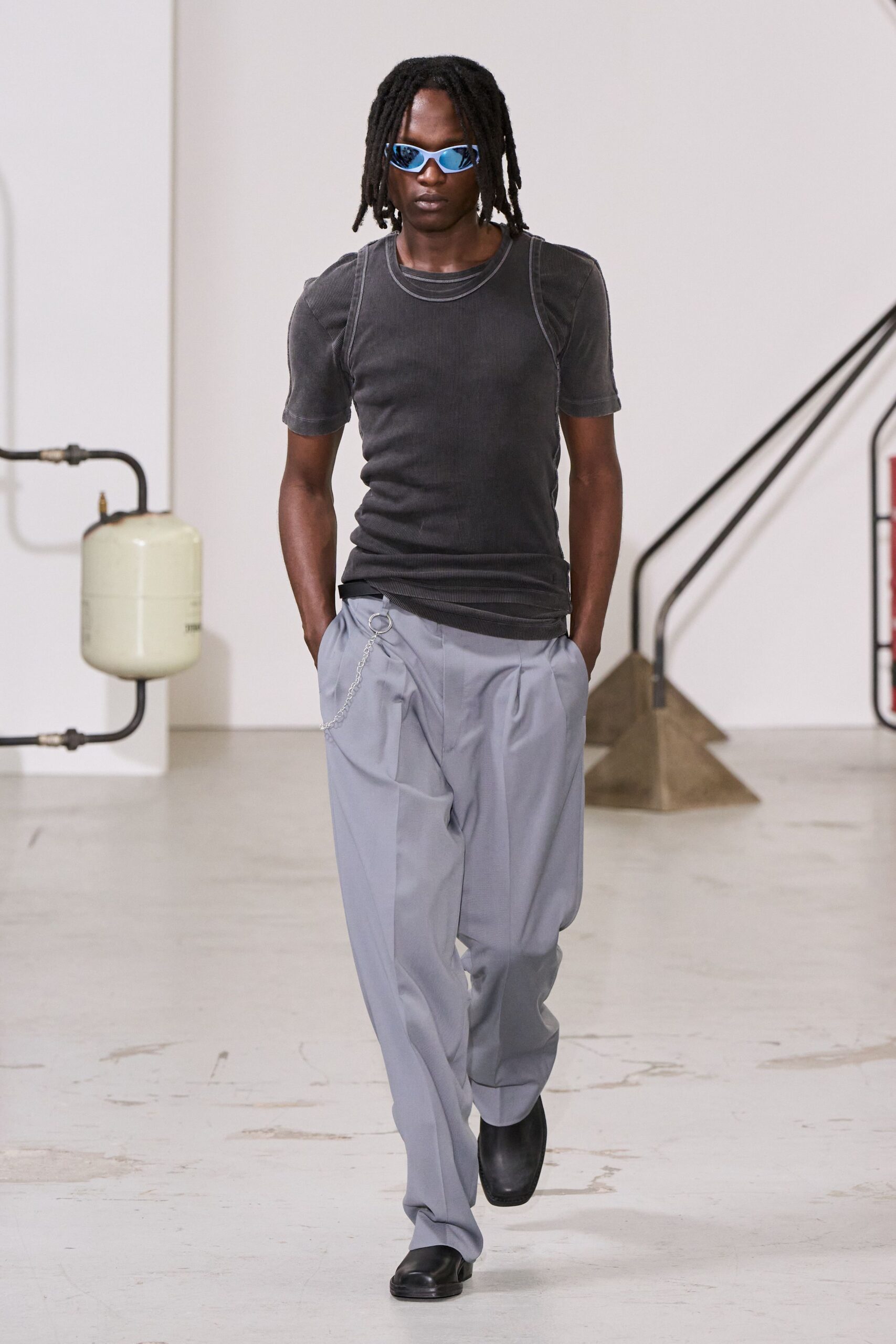 Wraparound shades with mirrored blue lenses. Washed black ribbed tee with a second neckline traced on top. Pale grey pleated trousers, silver wallet chain clipped at the hip. Square-toe black leather shoes. Walking across an empty office lobby before the air conditioning kicks in.