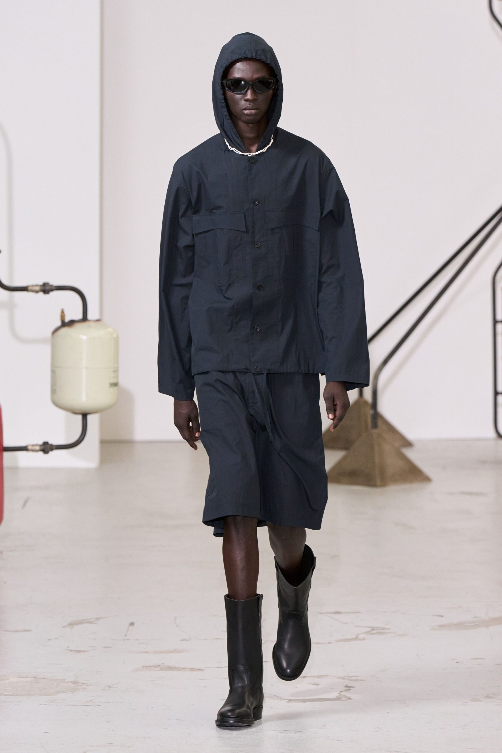Narrow wraparound black sunglasses. Ink cotton hooded shirt jacket with flap chest pockets and a rope-trim neckline. Matching long shorts in the same crisp cloth, hanging just below the knee. Matte black rubber boots with blunt rounded toes. Walking past industrial tanks after rain, keys damp in your pocket.