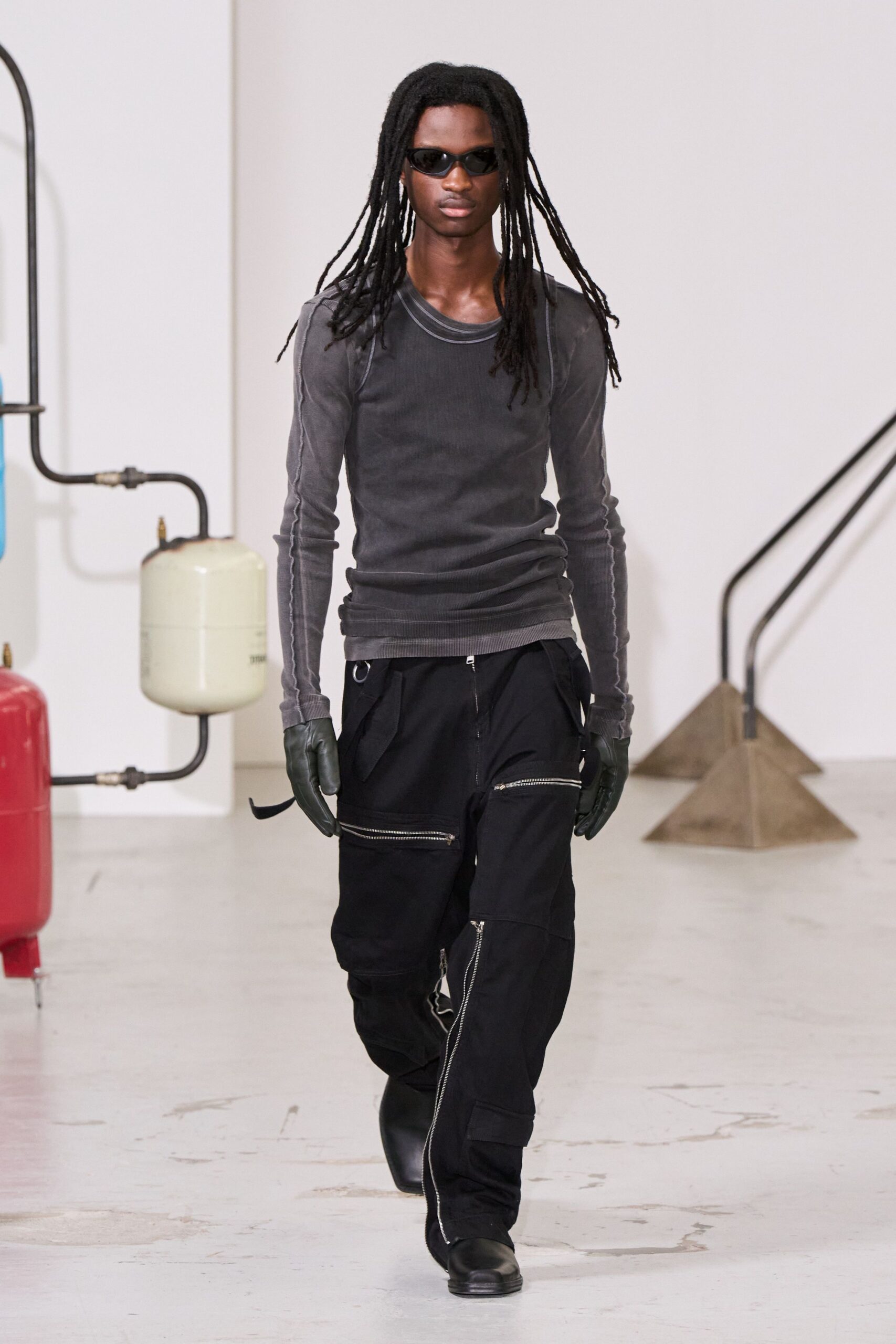 Narrow black wraparound frames. Washed charcoal ribbed long-sleeve, raw overlocked seams tracing the shoulders. Dark leather gloves. Black cargo trousers slashed with silver zips, one ring hanging at the waist. Square black boots. Walking the corridor outside a machine room after midnight maintenance.
