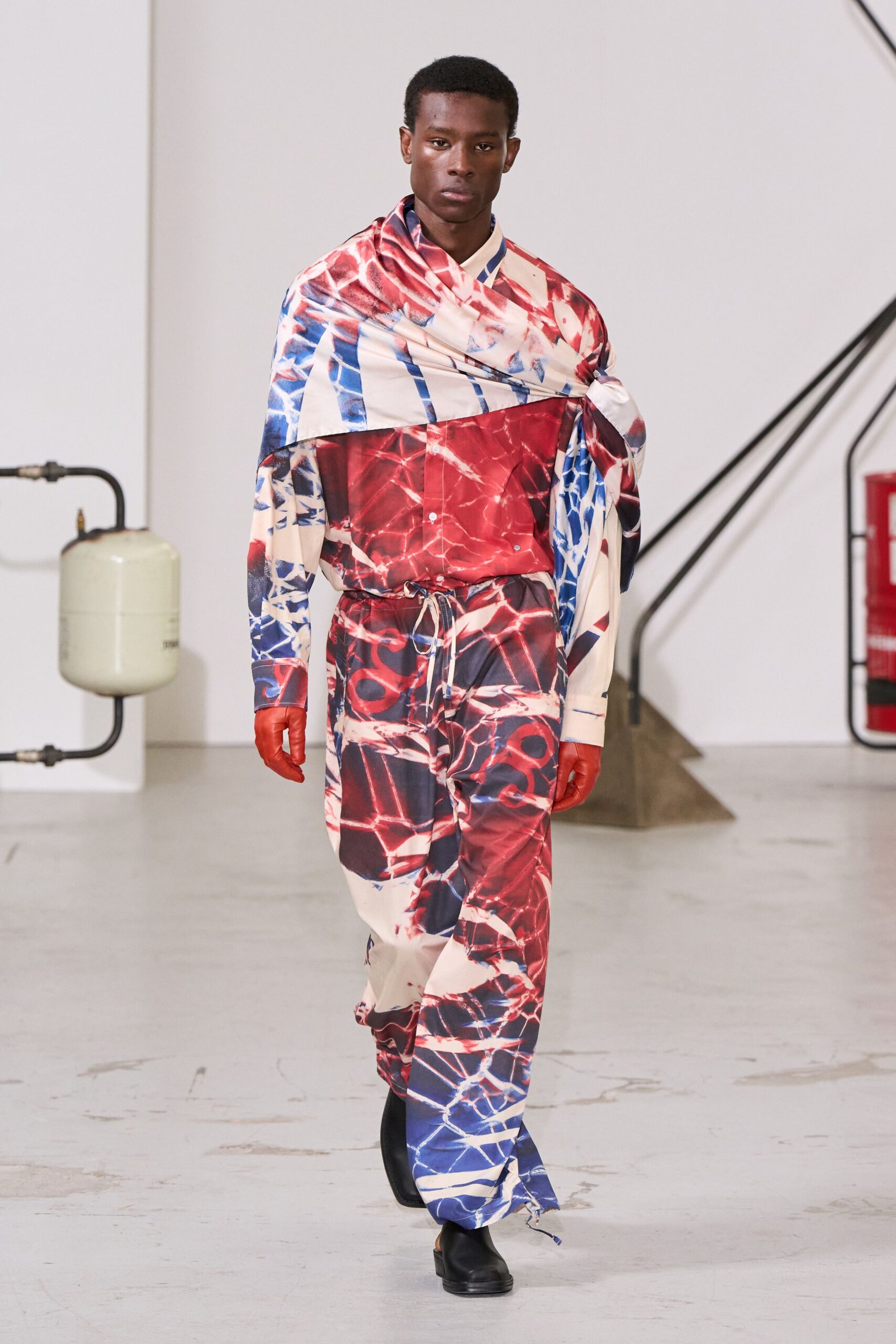 Oversized satin scarf in fractured red and cobalt, knotted at one shoulder. Matching print shirt with a pointed collar peeking through the wrap. Loose satin drawstring trousers, one cord end trailing by the shoe. Tomato leather gloves. Black square-toe clogs. Crossing an empty civic plaza after somebody set off the fountains.