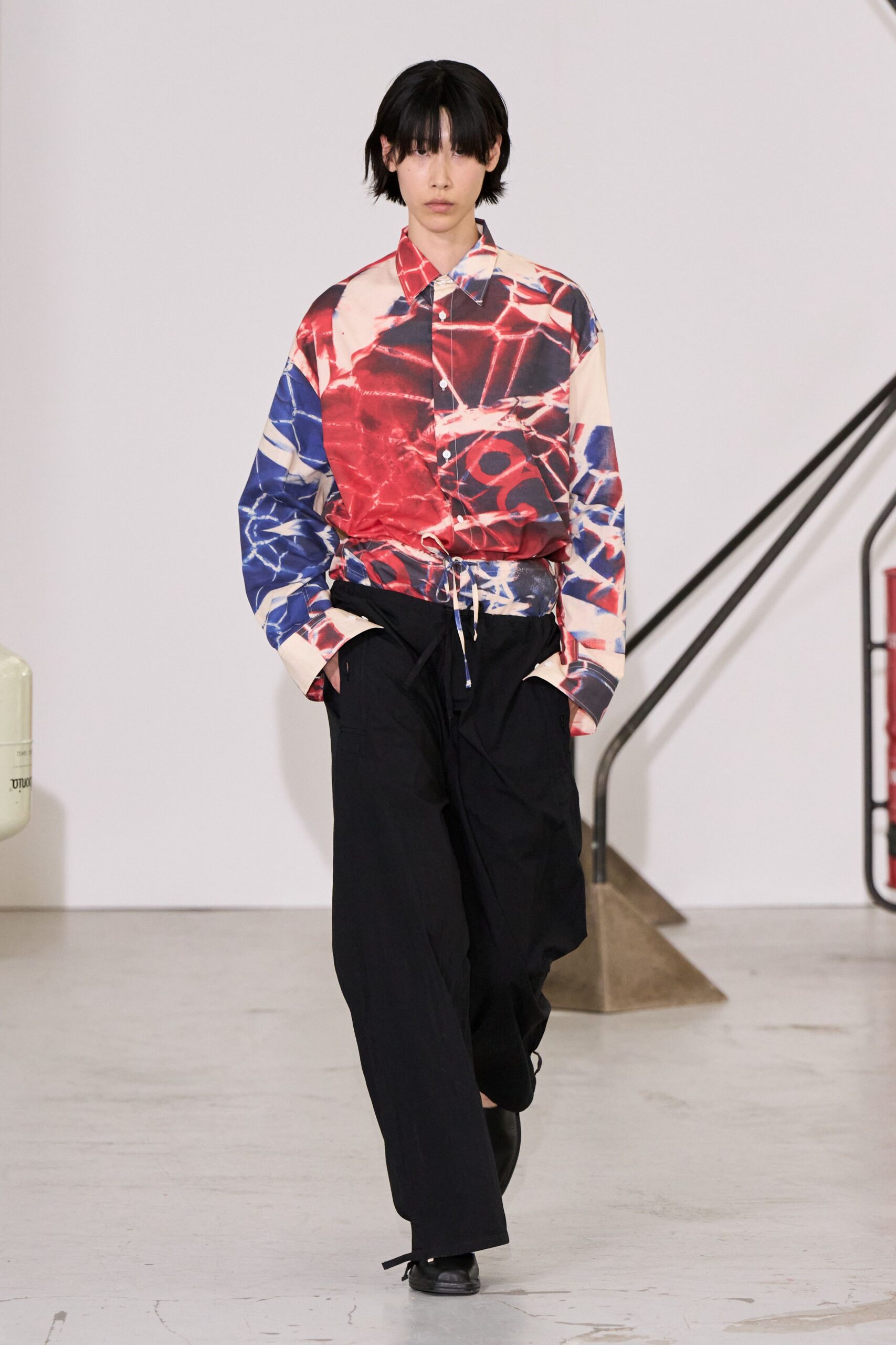 Oversized button-up printed in fractured crimson and cobalt, hem cinched with dangling cords. Black wide-leg cargo trousers in dry technical cloth, one ankle tie left loose. Square-toe black leather shoes. Walking into a municipal lobby where the heat has come on too early.