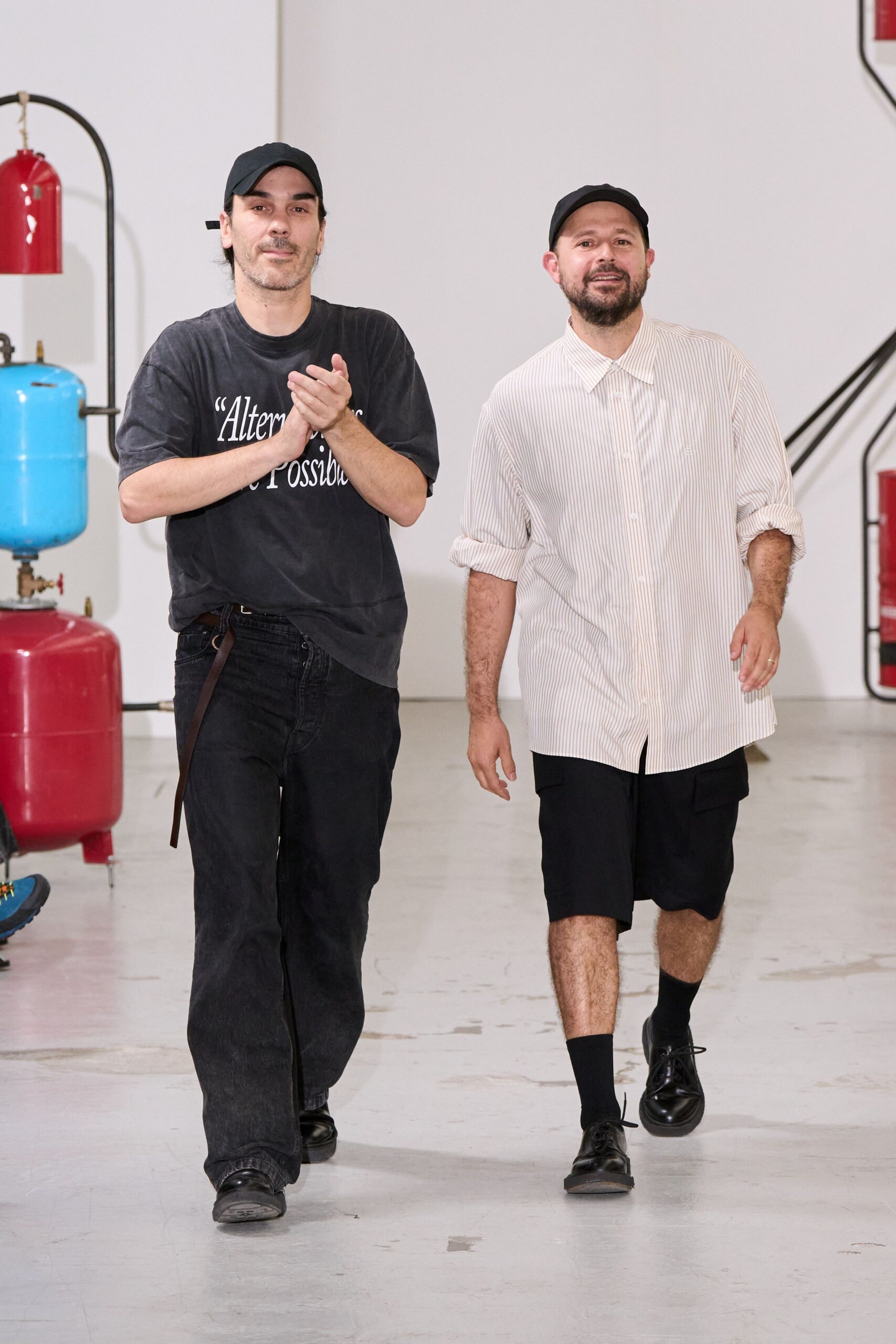 Black cotton baseball cap, brim curved low over the brow. White pinstripe button-up, sleeves rolled thickly to the forearms. Black knee-length shorts with a boxy, workwear cut. Plain black crew socks. Black leather derbies with one lace end trailing loose. Walking the factory floor after lunch, still clapping for the last pass.