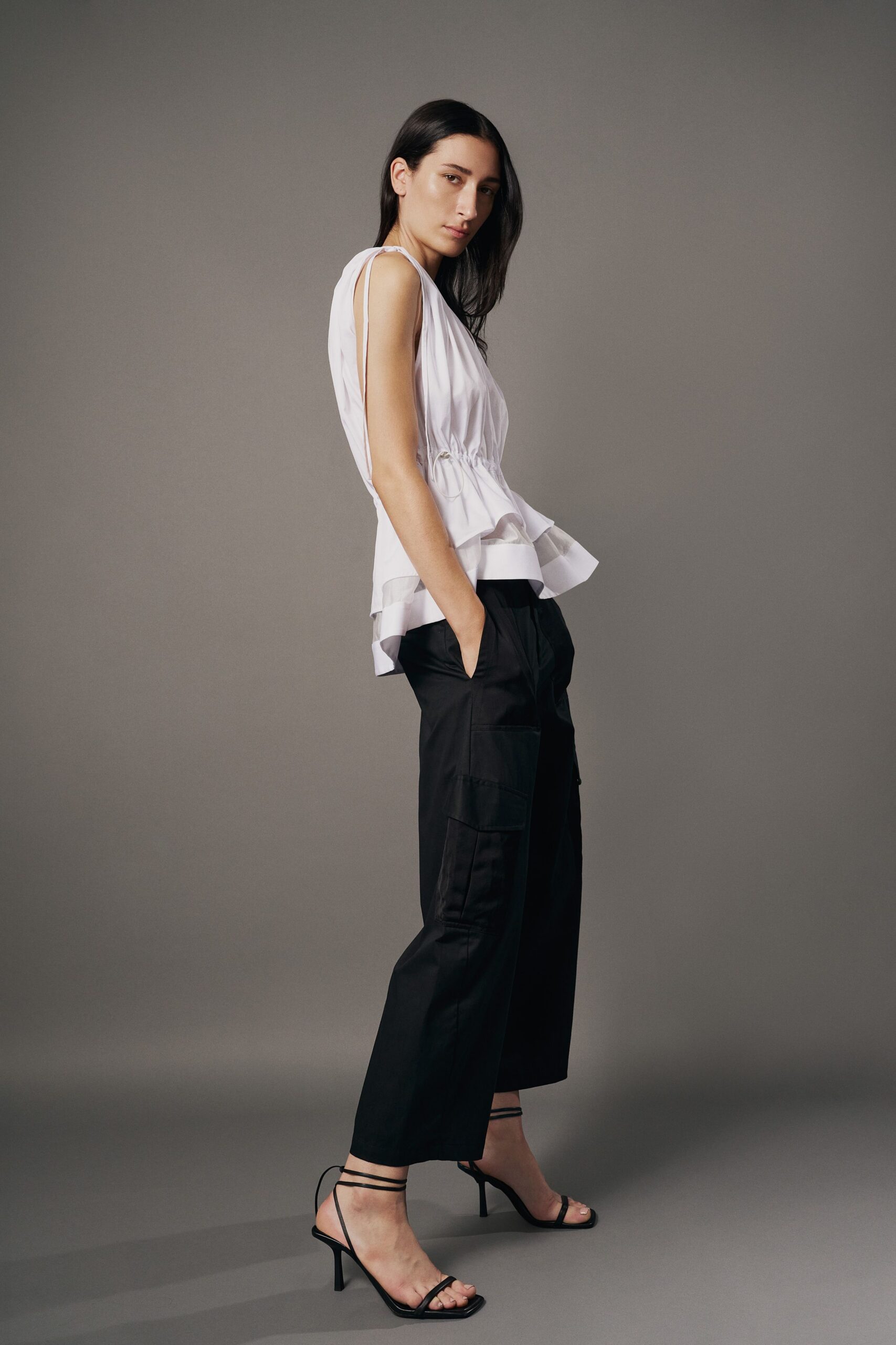 White sleeveless top with a gathered drawcord waist and sheer ruffled hem. Black cotton culottes, cropped wide leg with a single cargo patch pocket. Black barely there stiletto sandals with thin wraparound ankle straps. For the rooftop dinner where the wind keeps lifting the table linen.