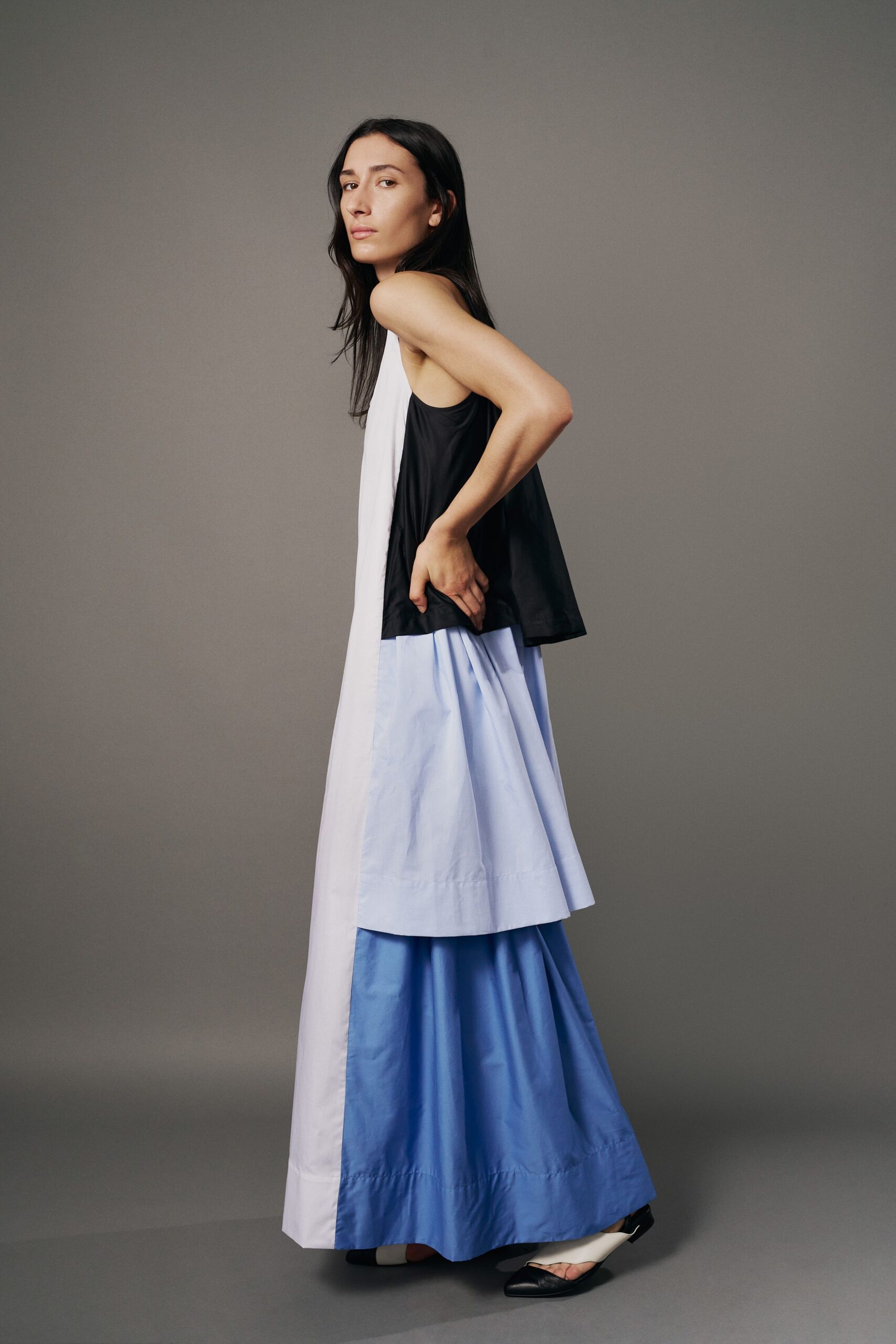 Apron-front column in white cotton, dropping into a sky-blue tiered hem. Black swing camisole overlay with a low armhole and crisp flare. Pointed black-and-cream mules with a curved cutout over the toes. Crossing a museum atrium before the air-conditioning fully wakes up.
