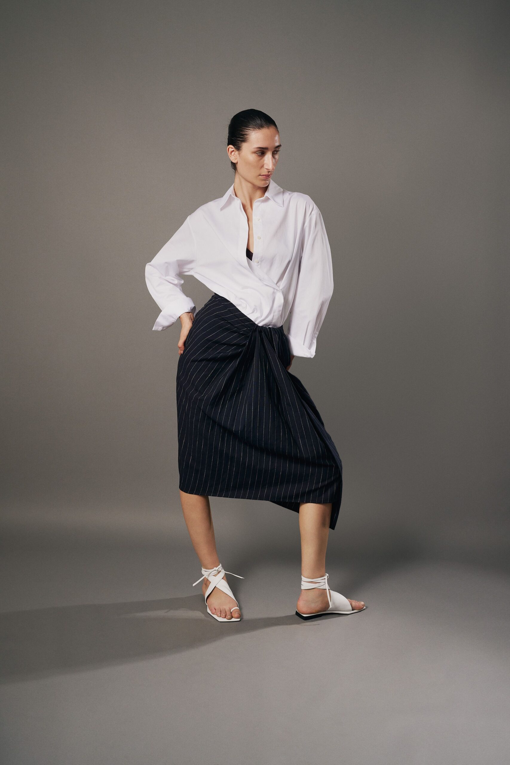 White cotton shirt bloused low, one cuff hanging wider than the other. Black triangle bralette peeking through the open placket. Navy pinstripe wrap skirt knotted into a heavy drape at the hip. White leather flats with thin laces winding the ankles. For the meeting where the fan is louder than anyone speaking.