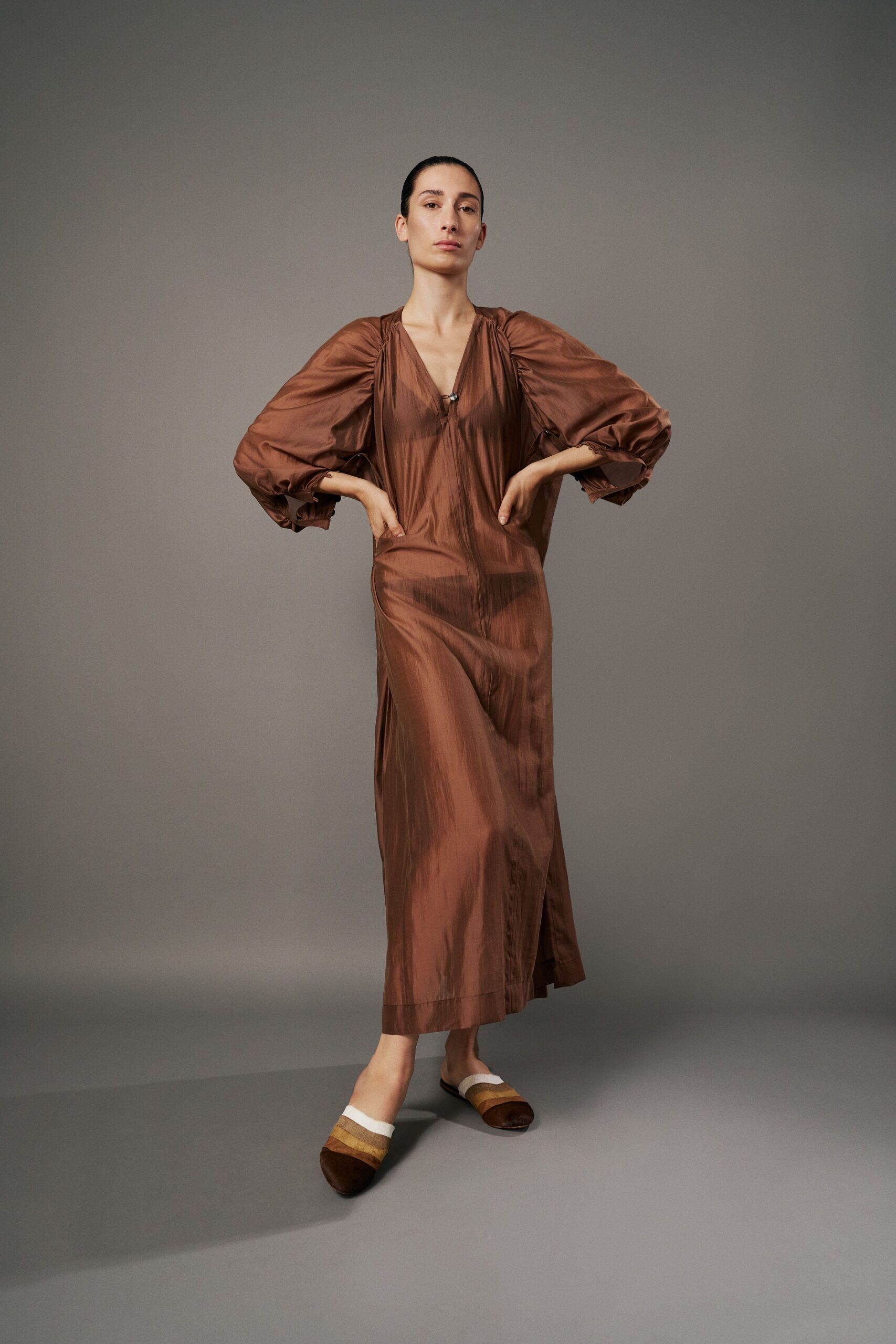 Coppery sheer dress in glazed silk, balloon sleeves gathered to narrow cuffs. Dark triangle bralette visible beneath the plunging V. Brown suede slides banded in camel and white stripes. Walking back from the pool bar with salt drying on your shoulders.
