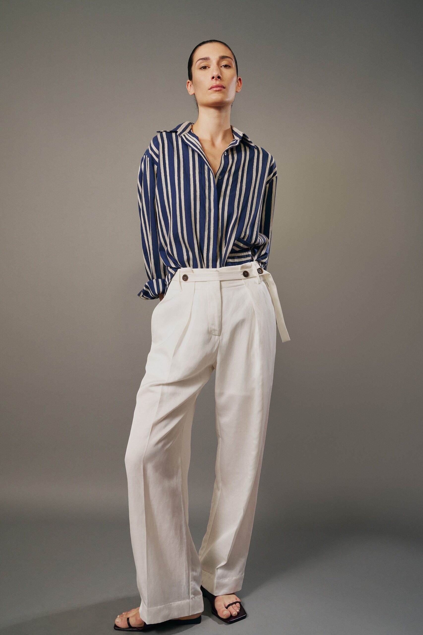 Blue and chalk striped shirt in airy weave, cuffs pushed and collar open. Ivory wide-leg trousers with double waistband buttons and a loose self belt. Black square-toe thong sandals. For checking into a whitewashed hotel before the room is ready.