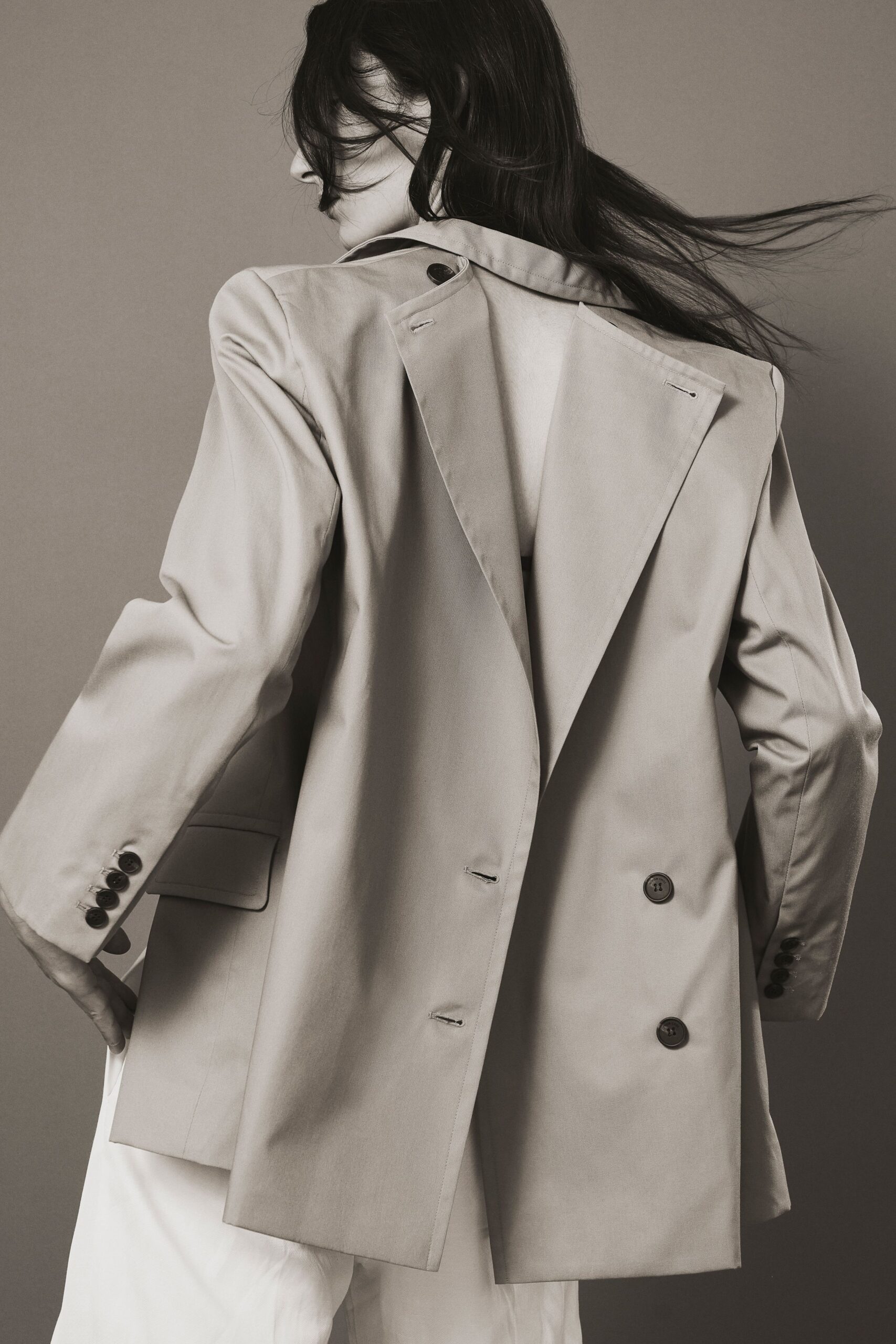 Stone double-breasted blazer with a split back panel hanging loose from the shoulder button. White trousers in a fluid, lightly wrinkled cloth. Wind off the ferry, turning around before anyone says your name.