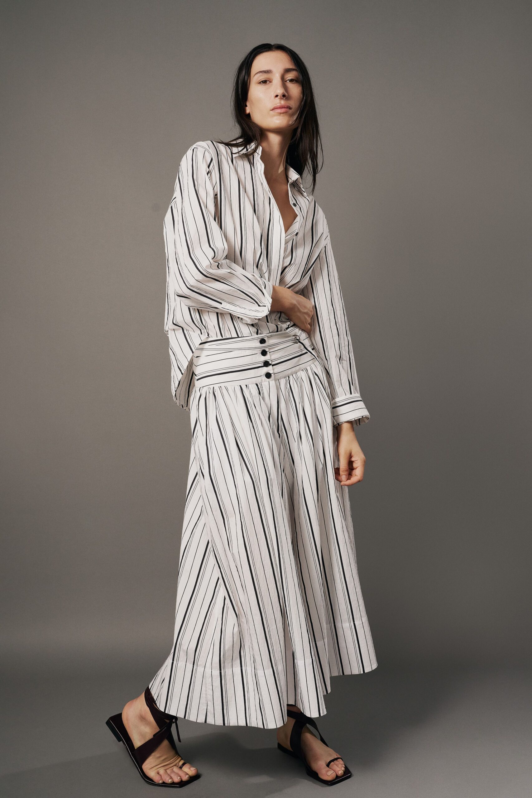 White striped shirt in airy cotton, worn open and slightly askew. Matching gathered midi skirt with a broad waistband and four black buttons. Black thong sandals with square toes and slim ankle ties. Windows open before noon, hotel sheets still creased behind you.