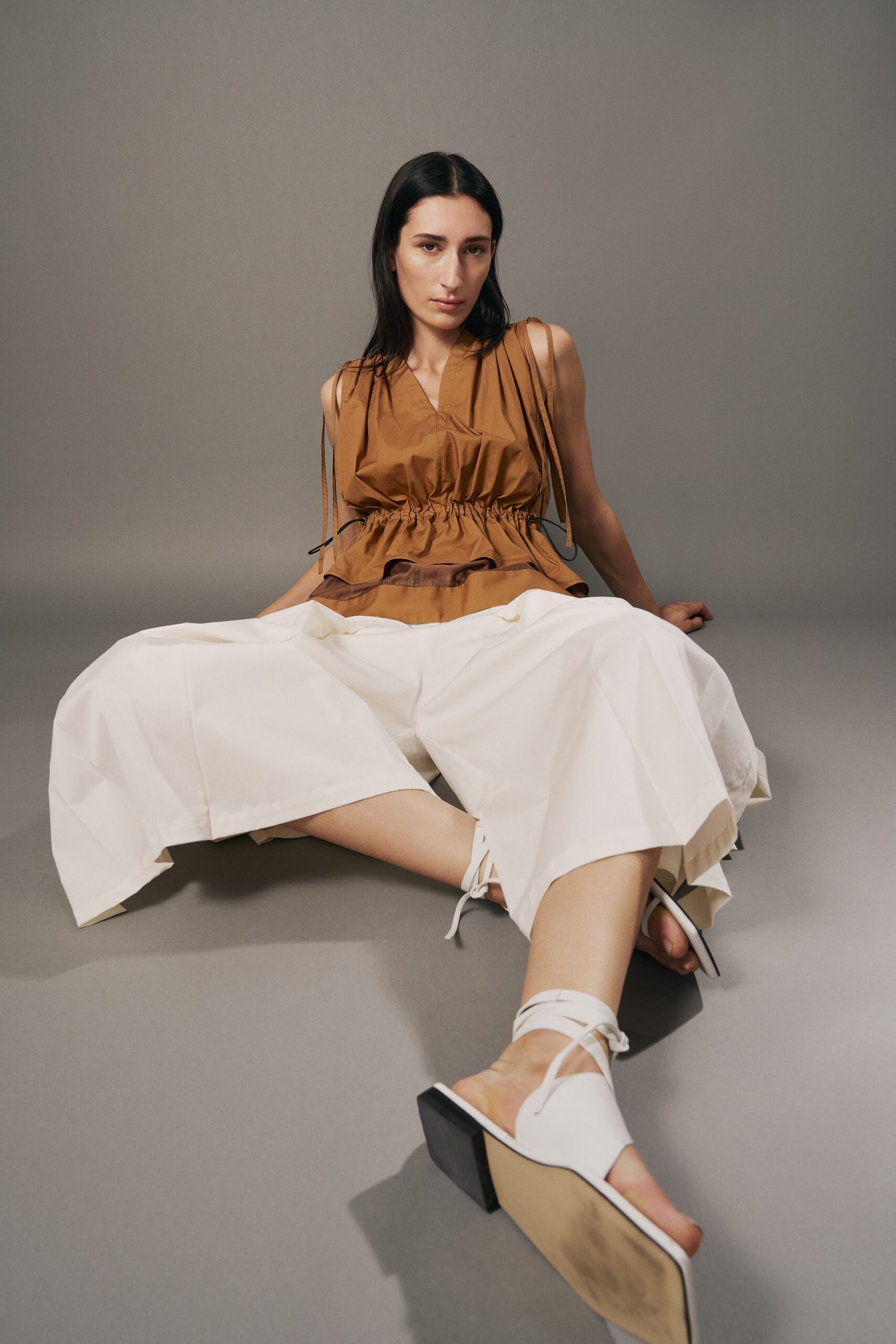 Tobacco poplin top with ruched drawstring waist and ribbon shoulder ties. Crisp white culottes cut extra wide, creased into angular planes. White lace-up mules with square toes and pale wood wedge soles. Sitting on the cool gallery floor after the installers have gone.