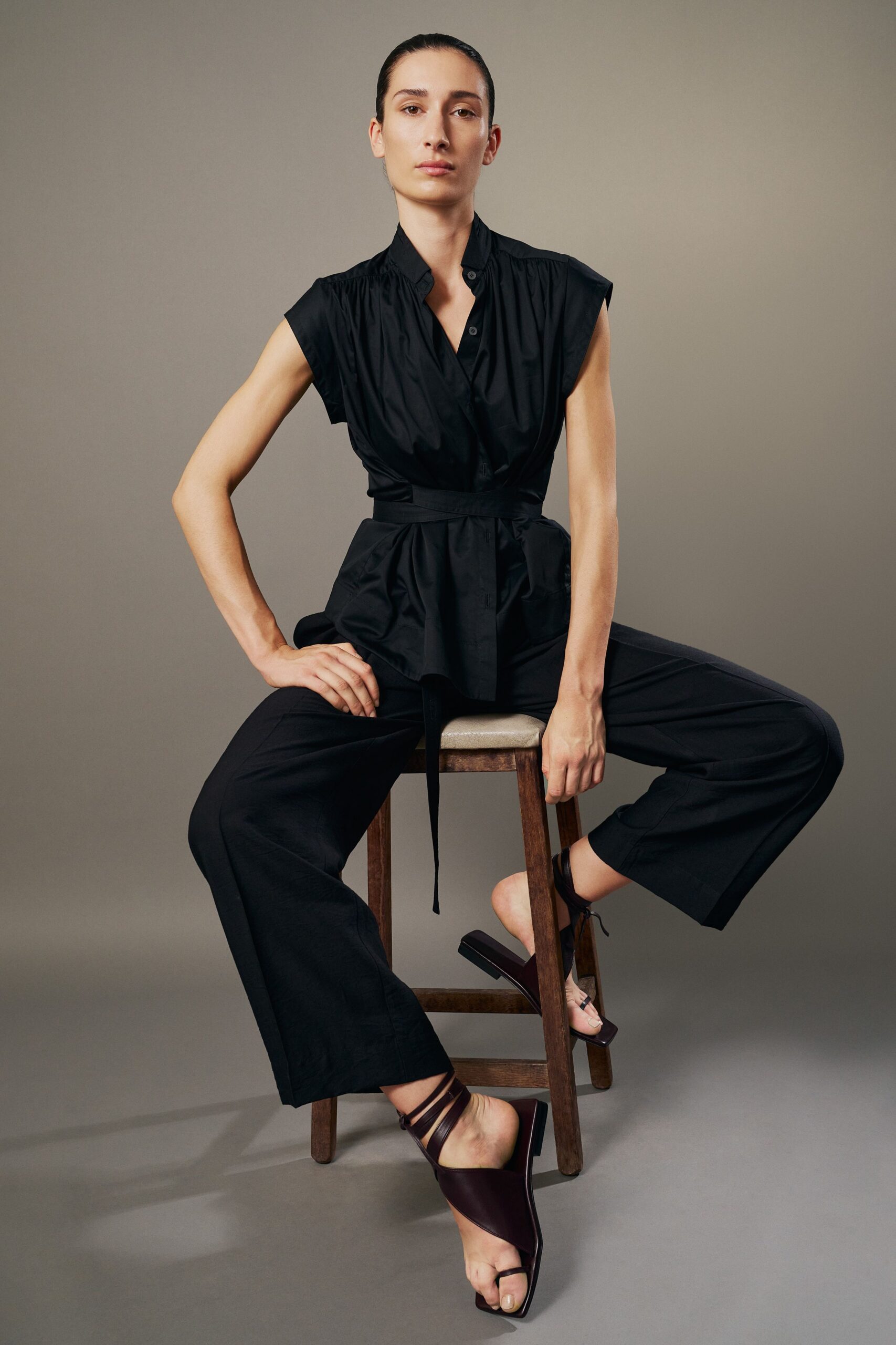 Black cotton blouse with cap sleeves, gathered neckline and long self-tie belt. Black straight trousers in crisp plain weave, hem breaking clean at the ankle. Oxblood leather thong sandals with narrow straps winding the ankle. For the courtyard dinner after sunset when the stone still holds heat.