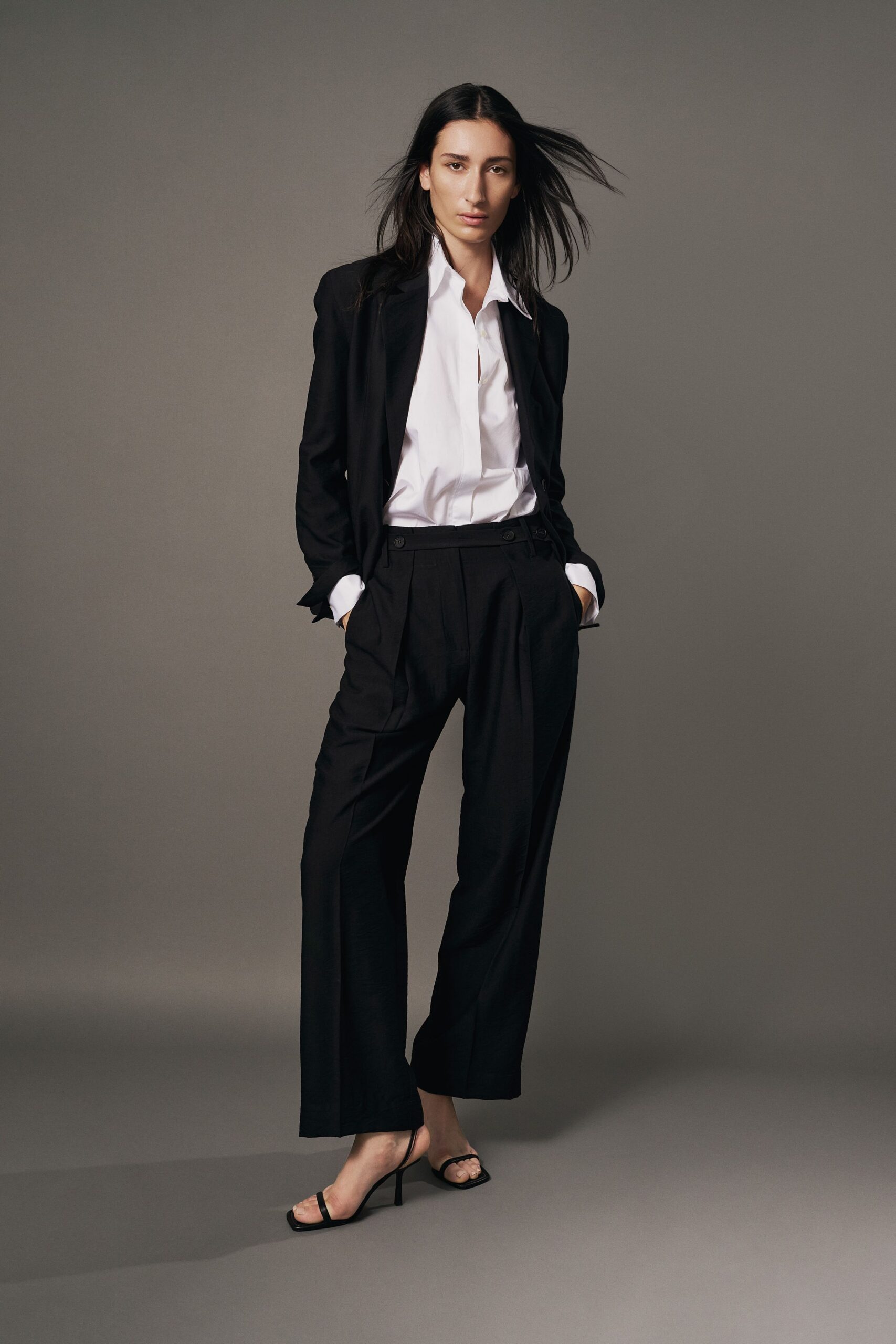 Black wool blazer cut lean through the body, sleeves pushed long. White poplin shirt left open at the throat, cuffs turned back. Black pleated trousers with side-tab buttons and a deep dropped rise. Barely there black thong heels. For the meeting where you arrive calm and leave with the lease.