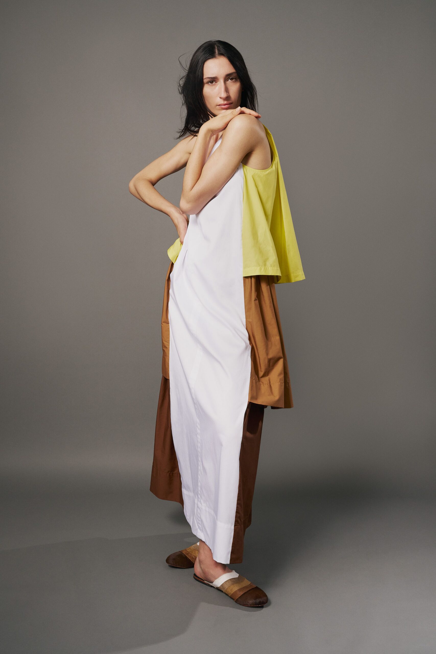 White poplin column dress, side seam pulling slightly across the hip. Citron cotton overlay falling loose from the shoulder blades. Caramel wide trousers peeking below, crisp with a low stitched cuff. Flat mules in sand and tobacco bands. Walking back from a hot courtyard lunch, napkin still in your lap.
