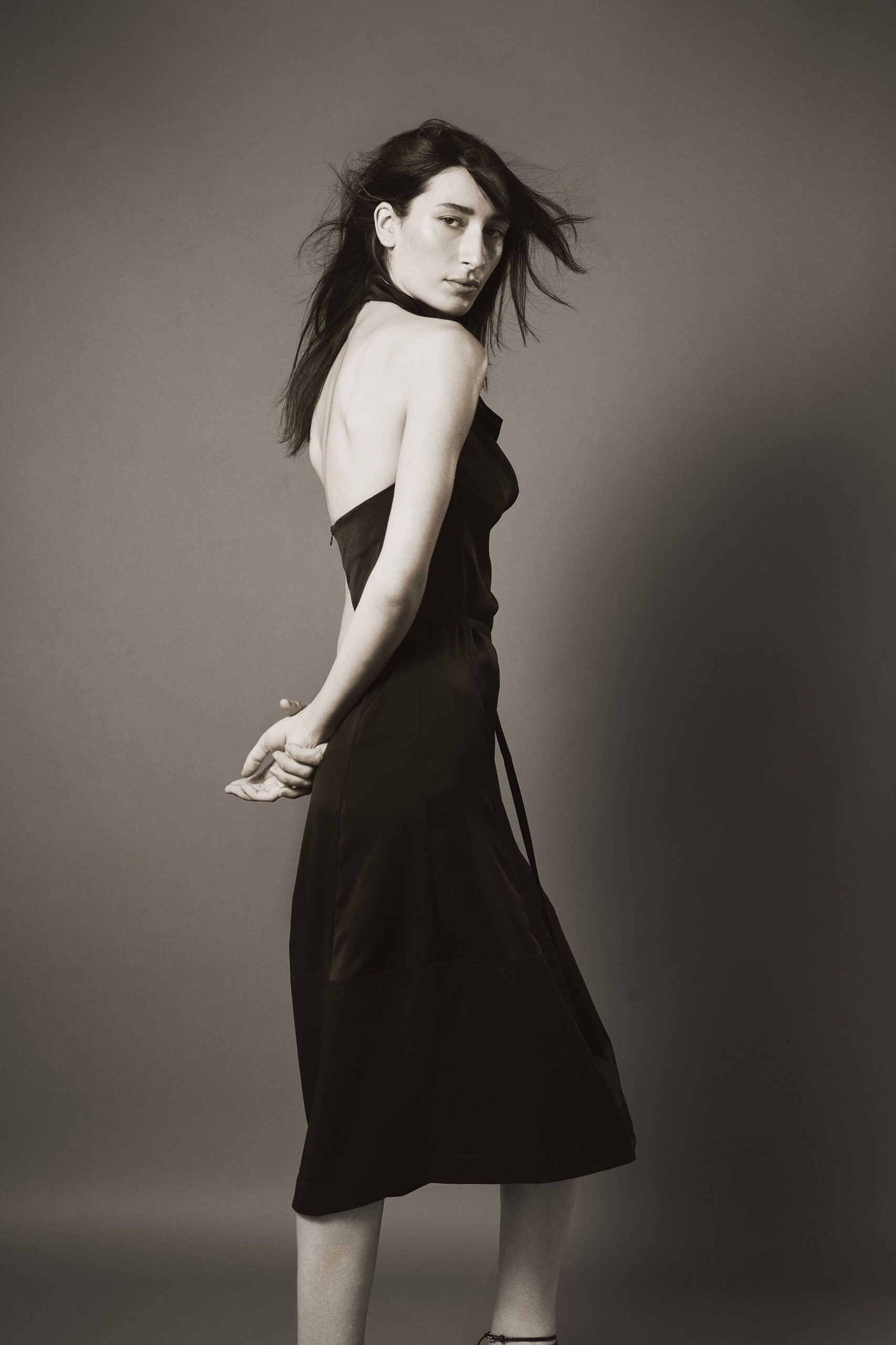 Black halter dress in matte sateen, back cut low with a dangling waist tie. Crossing the hotel corridor barefoot after the wedding finally ended.