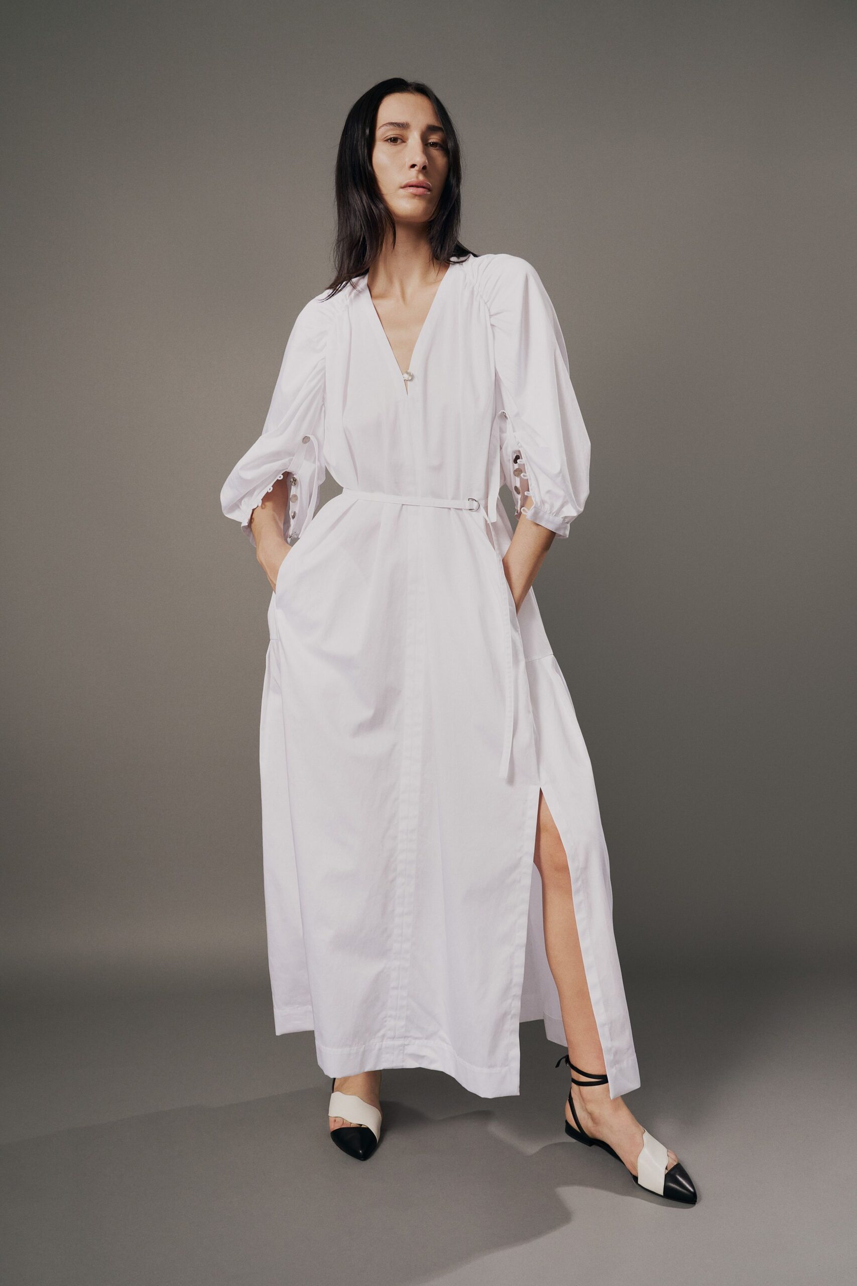 White cotton-poplin midi dress with gathered shoulders, side-buttoned sleeve openings and a thigh slit. Narrow self belt cinched through a small silver D-ring. White flats with black cap toes and thin wraparound ankle ties. For opening the shutters in a rented house before the heat gets in.