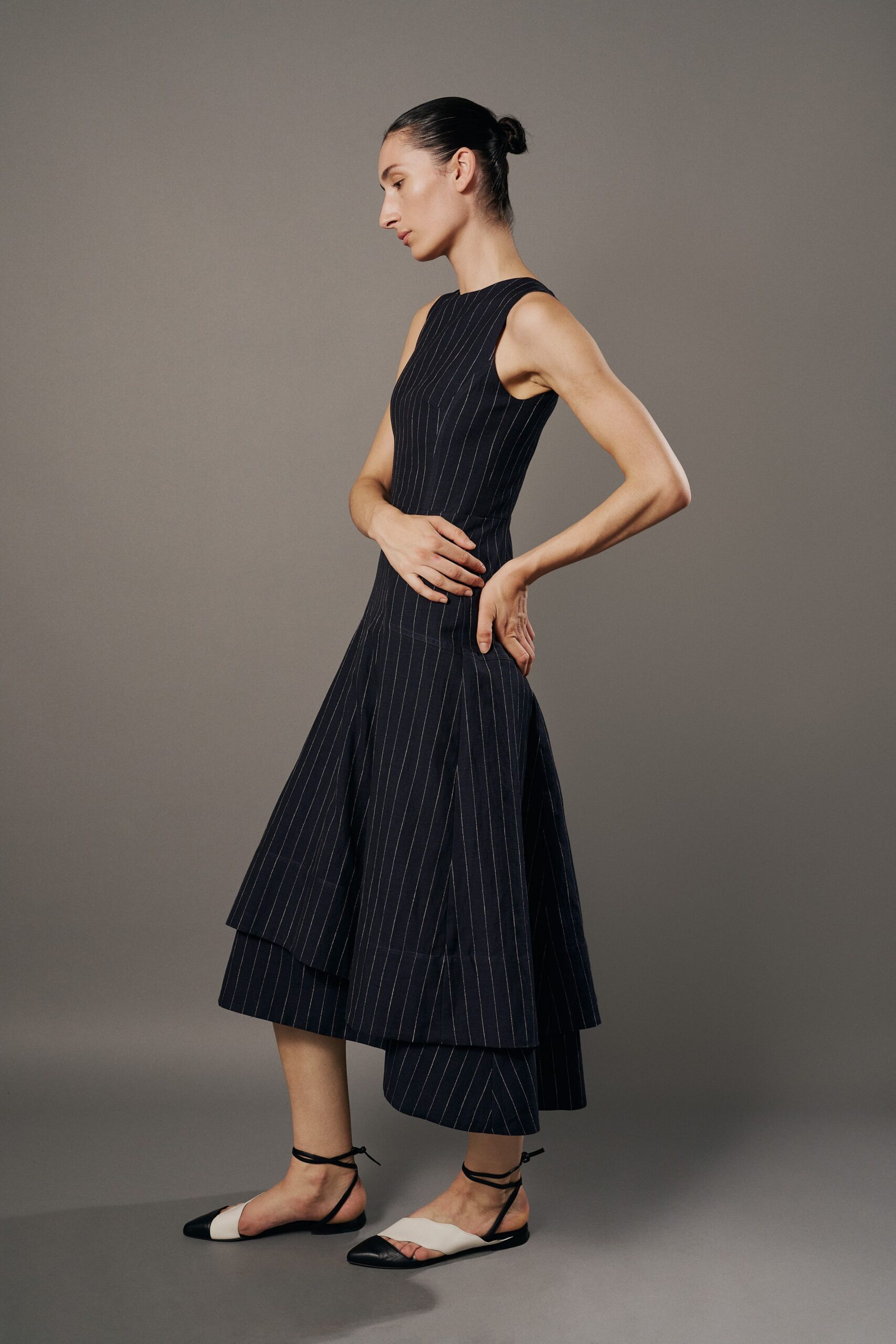 Navy pinstripe dress with a close crew neck and layered handkerchief panels. Black-and-ivory slingback flats tied with skinny wraparound ankle cords. Walking into a municipal atrium for someone else's architecture lecture.