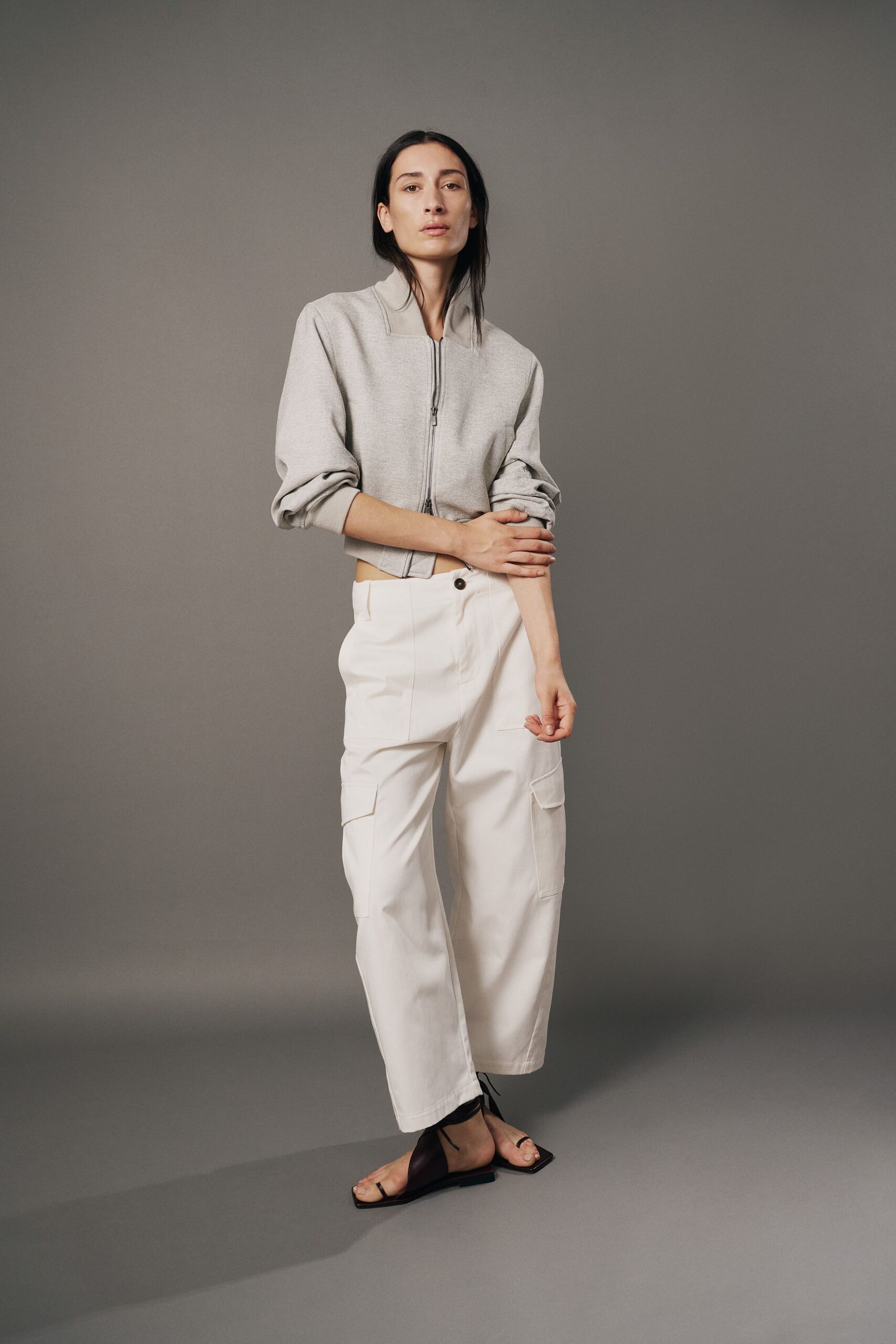 Pale grey knit zip jacket, cropped hem tipping up at the front. Chalk cargo trousers with broad patch pockets and a dark single button. Black square-toe thong sandals, ties wrapped once at the ankle. Walking across a museum courtyard before the paving gives off heat.