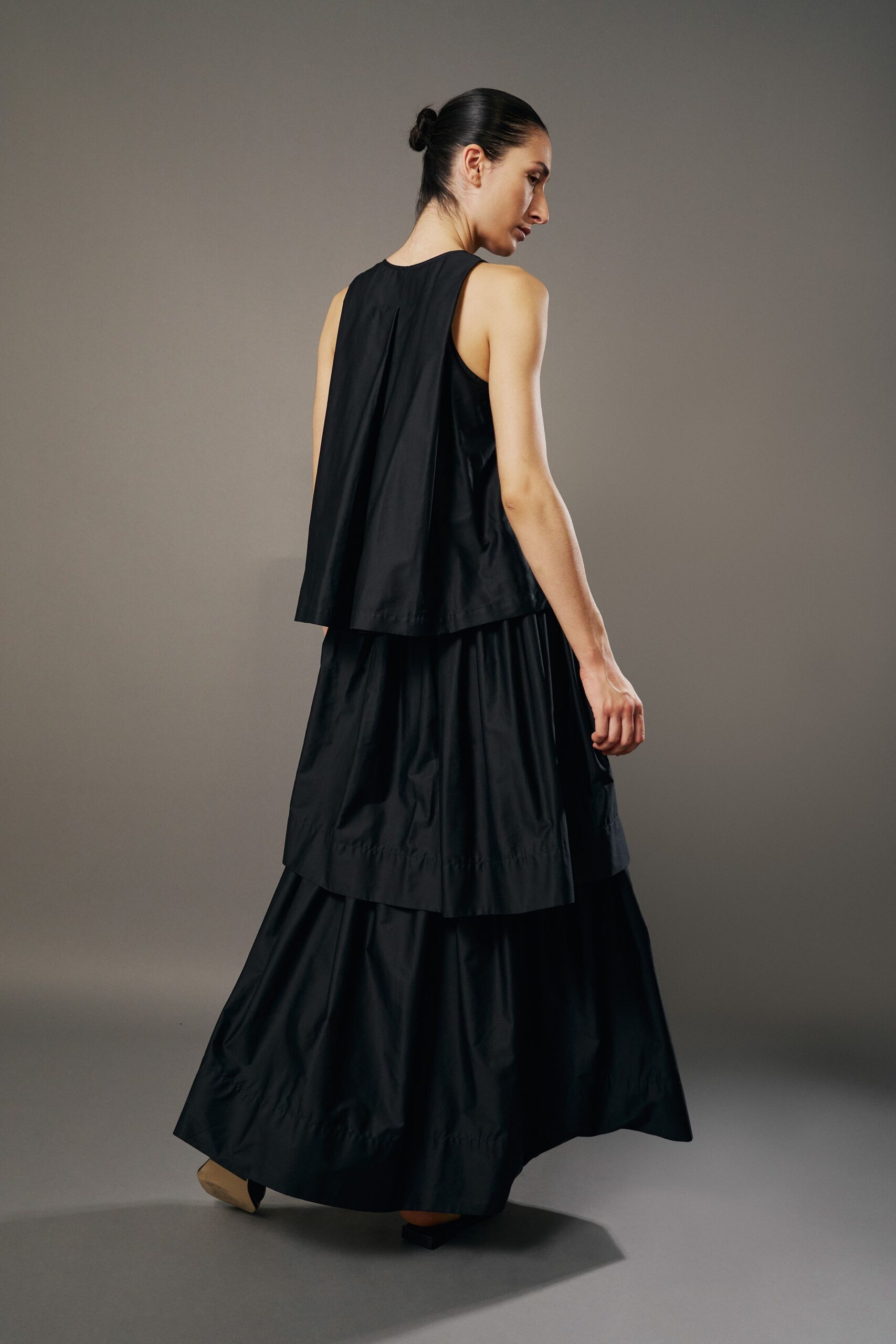 Black cotton poplin shell, back pleat opening into a cape-like panel. Matching tiered maxi skirt with a dropped seam circling the lower calf. Tan square-toe flats peeking under the hem. Crossing a museum courtyard just before the doors lock for rain.